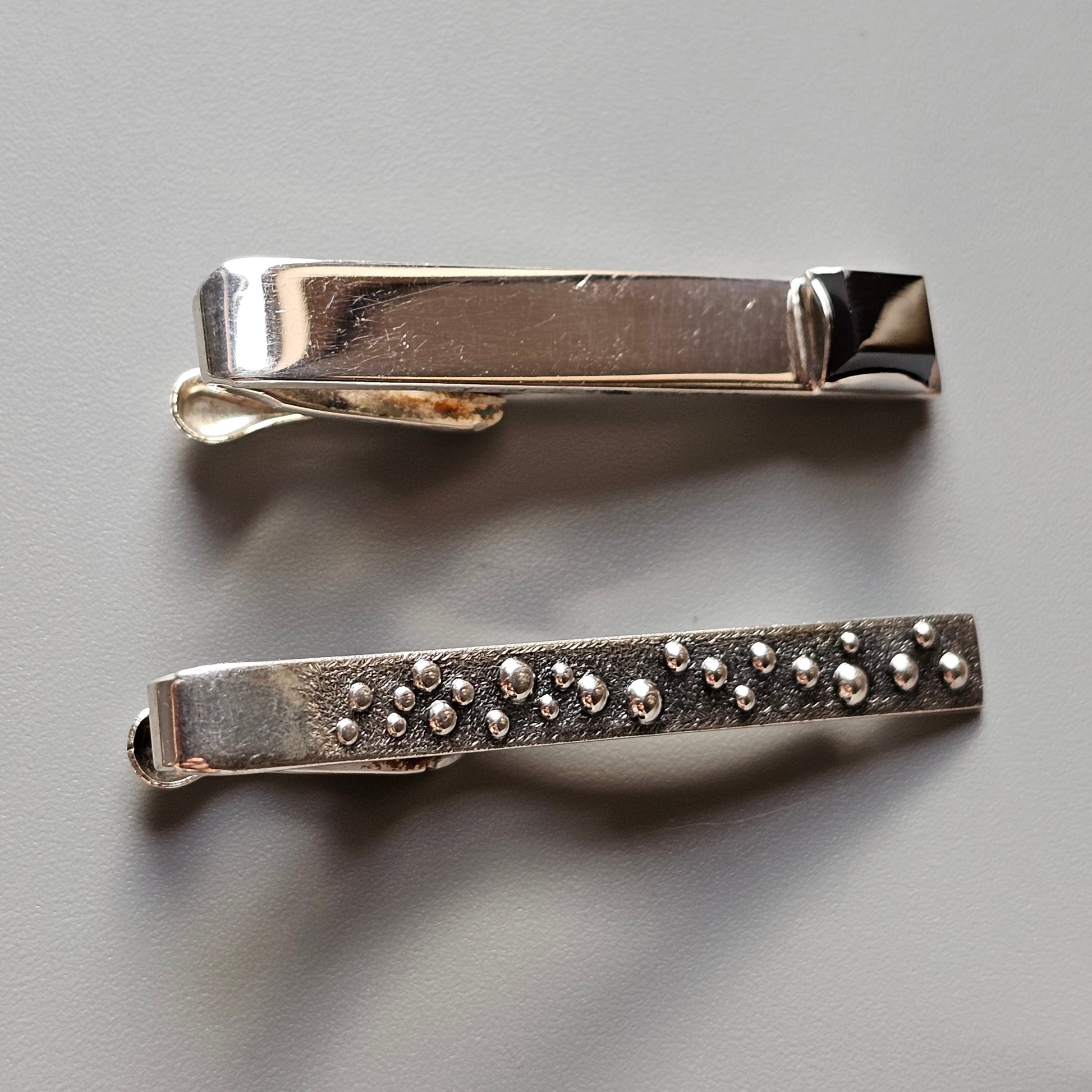 Two metal tie clips, one plain and one decorated with small studs.