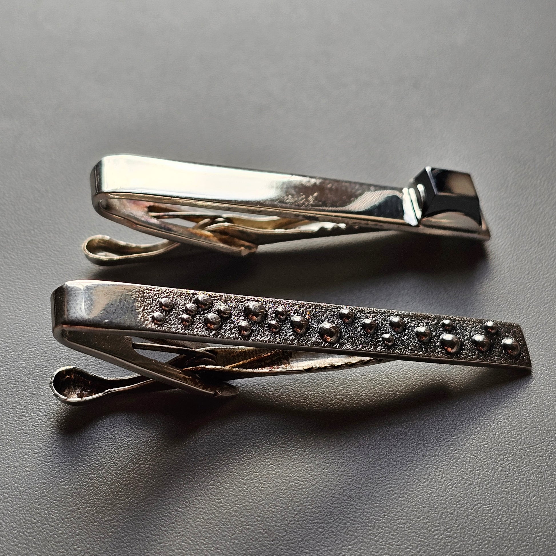 Two metal tie clips, one plain and one decorated with small stones or studs.