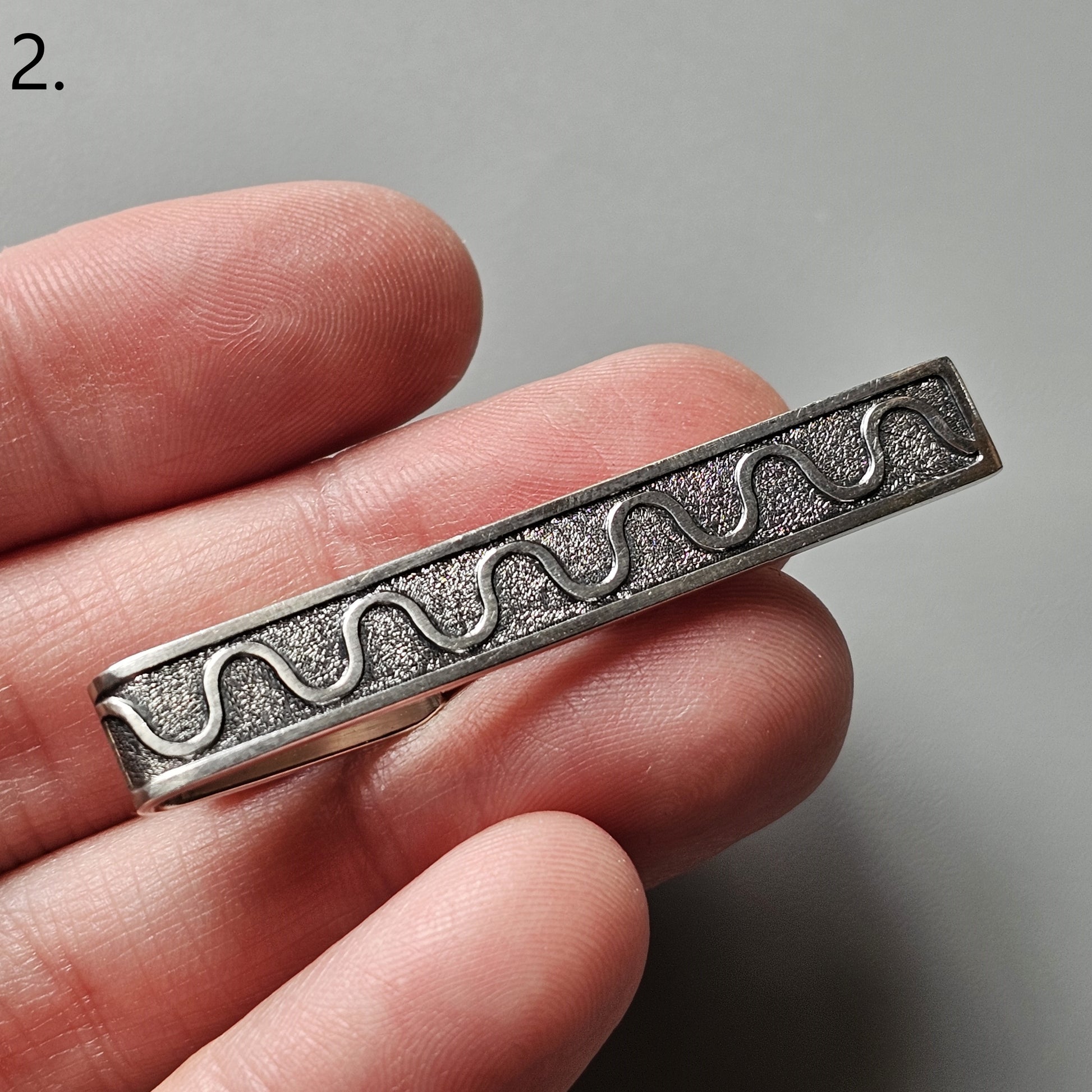 Metal tie clip or bar with a wavy pattern engraved on its surface.