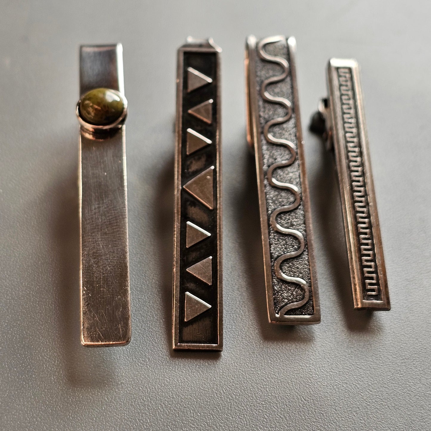 Four decorative metal tie clips with different engraved patterns.