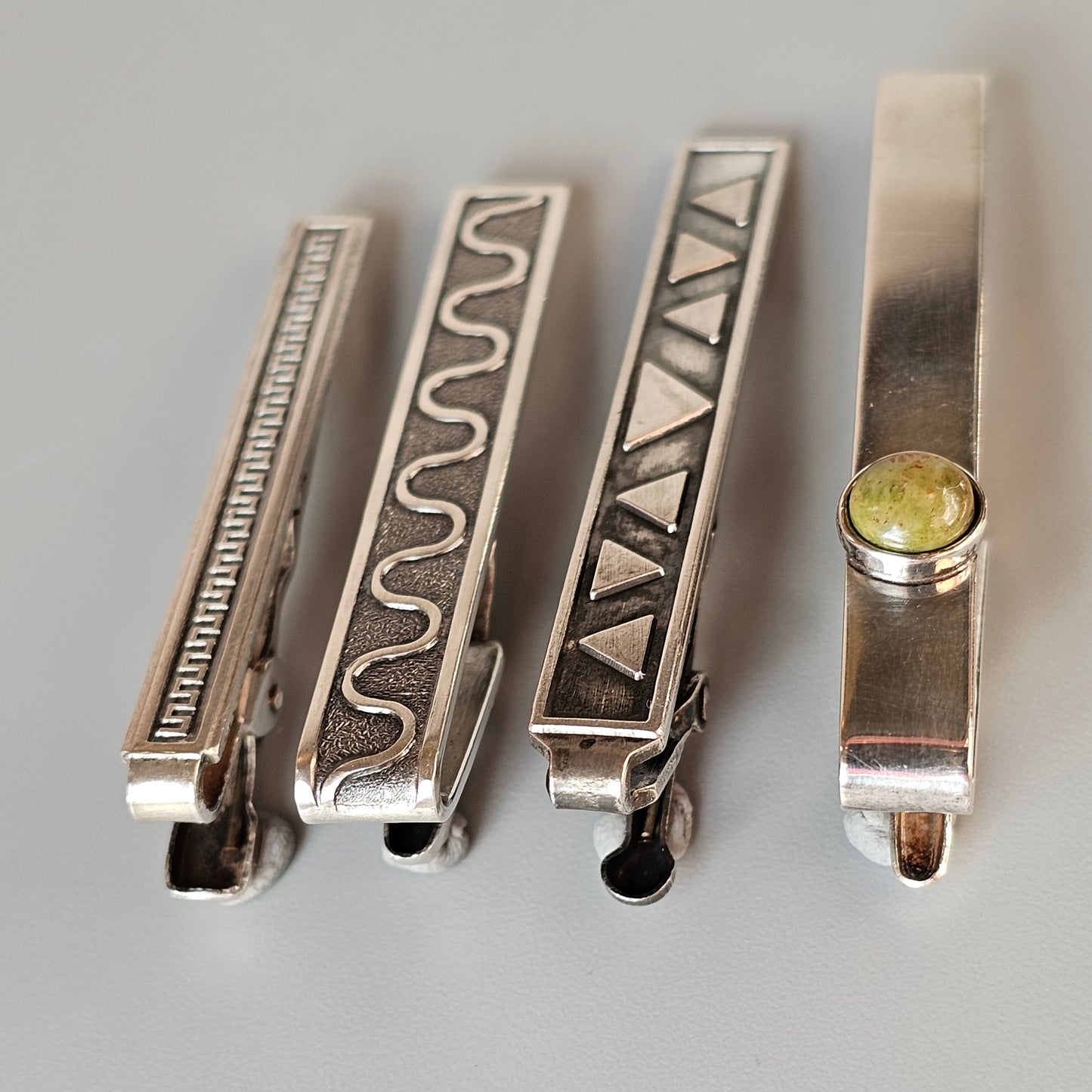 Four decorative metal tie clips with different engraved patterns.