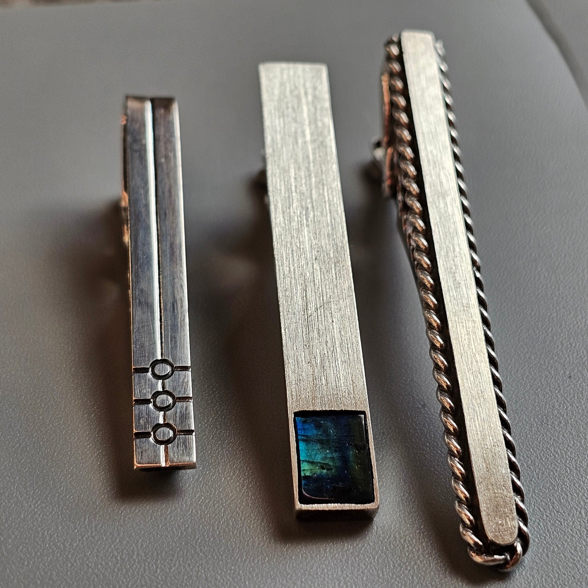 Three metallic tie clips with different designs and decorative elements.