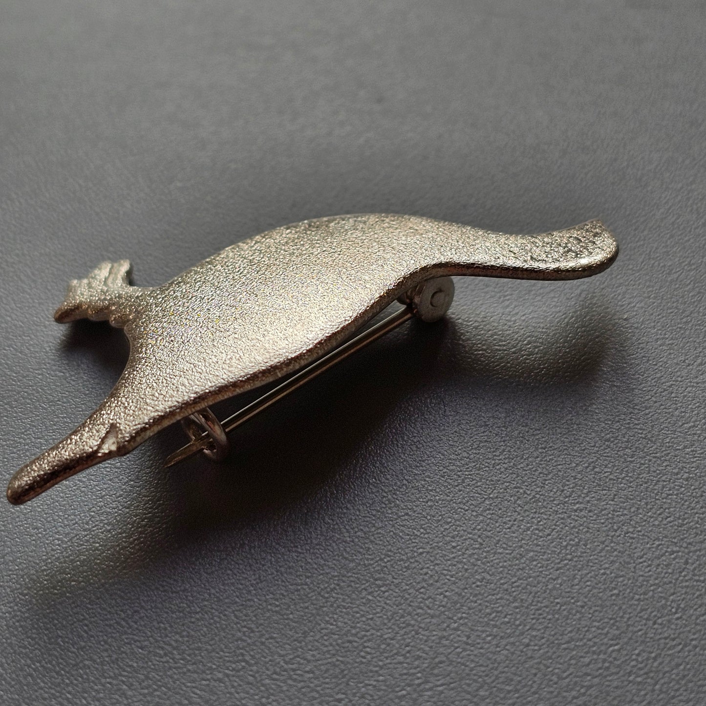 Metallic bird-shaped brooch or pin with an elongated body and spread wings.