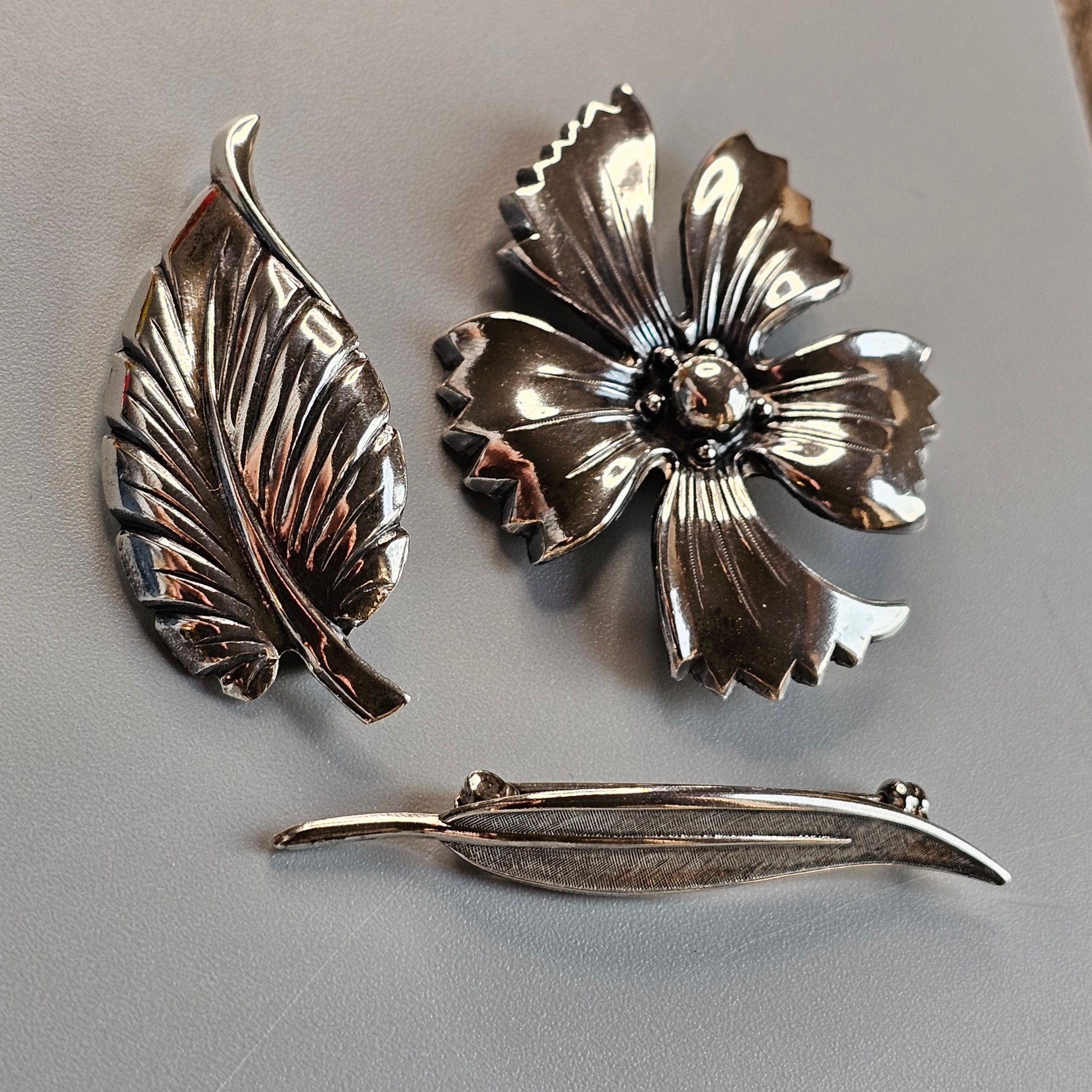 Three silver-toned metal brooches shaped like botanical elements: a leaf, a flower, and a feather.