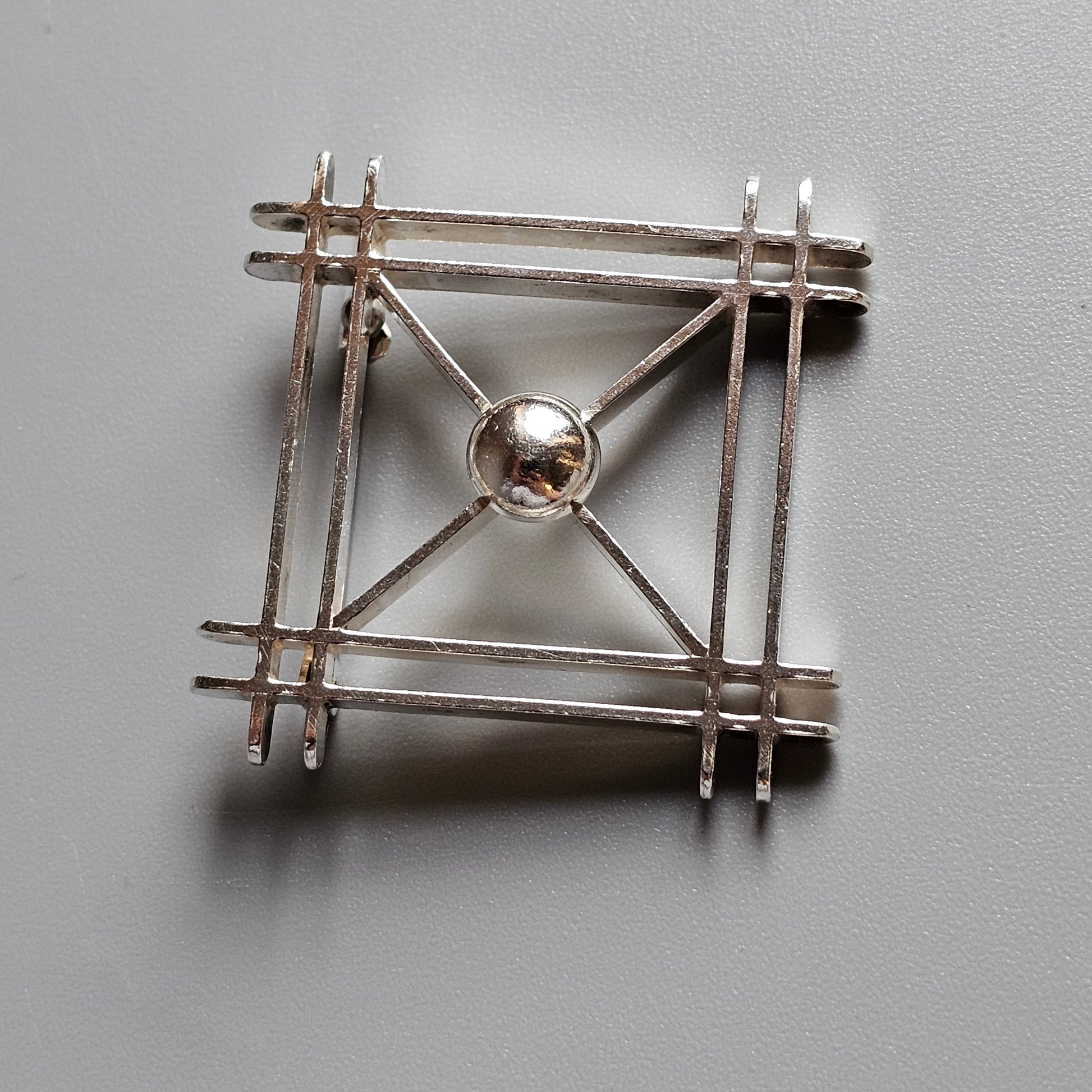 Square-shaped silver brooch with intersecting bars and a central spherical bead.