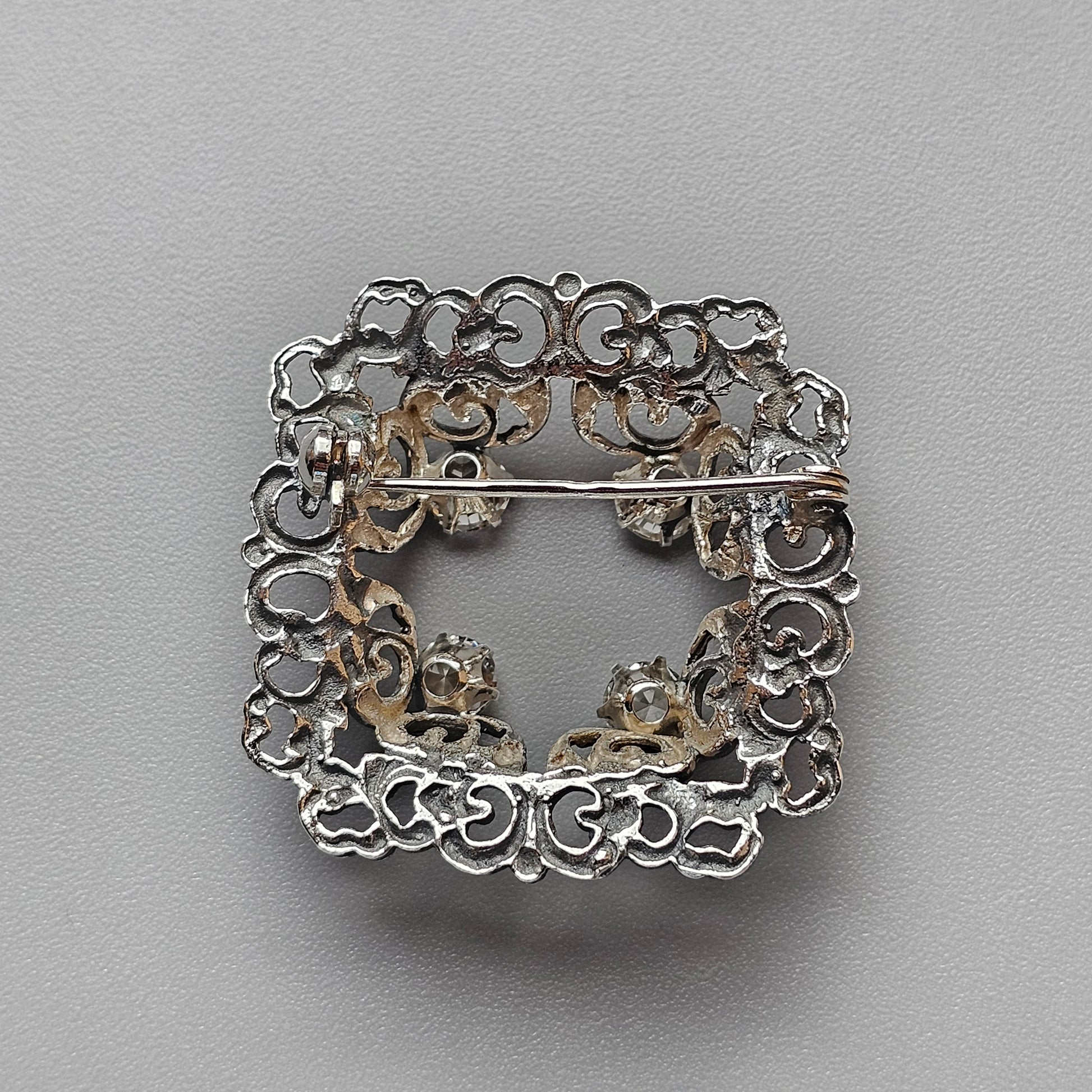 Ornate silver brooch with intricate filigree scrollwork forming a square shape.