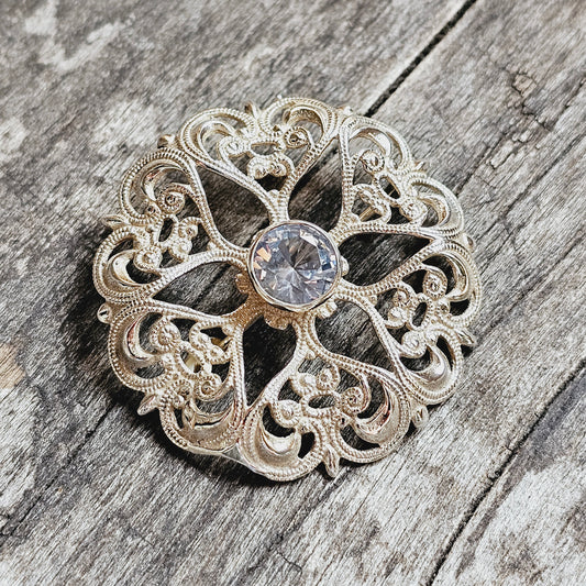 Ornate silver brooch with intricate filigree design and central clear gemstone.