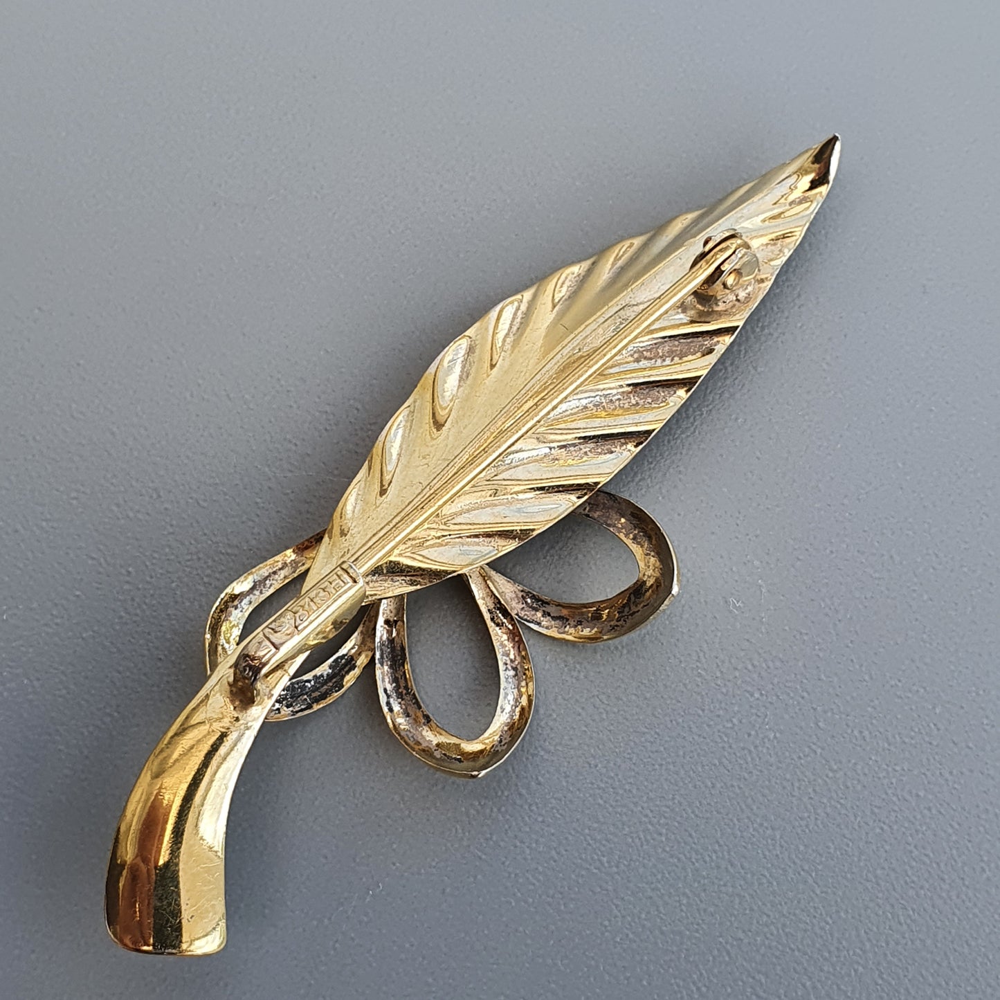 Gold-colored feather-shaped brooch or pin with a decorative bow design.