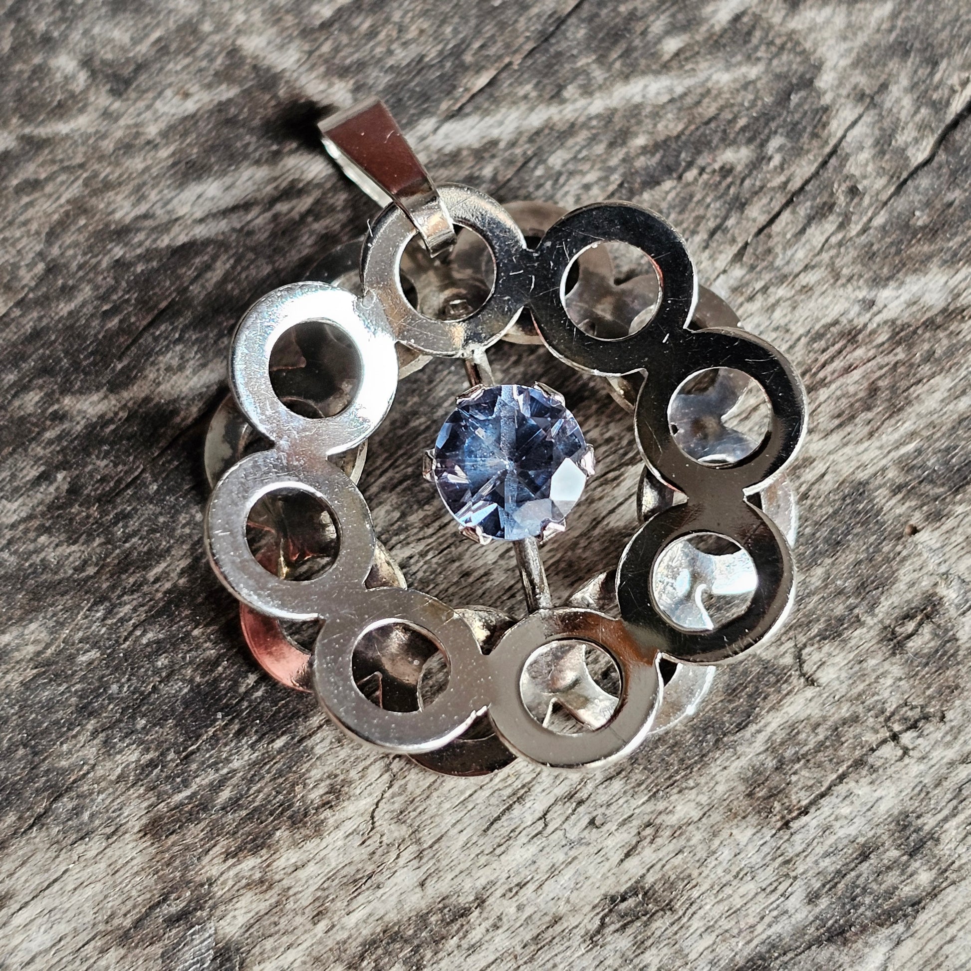 Circular metal pendant with a blue gemstone at its center, surrounded by ring-shaped elements.