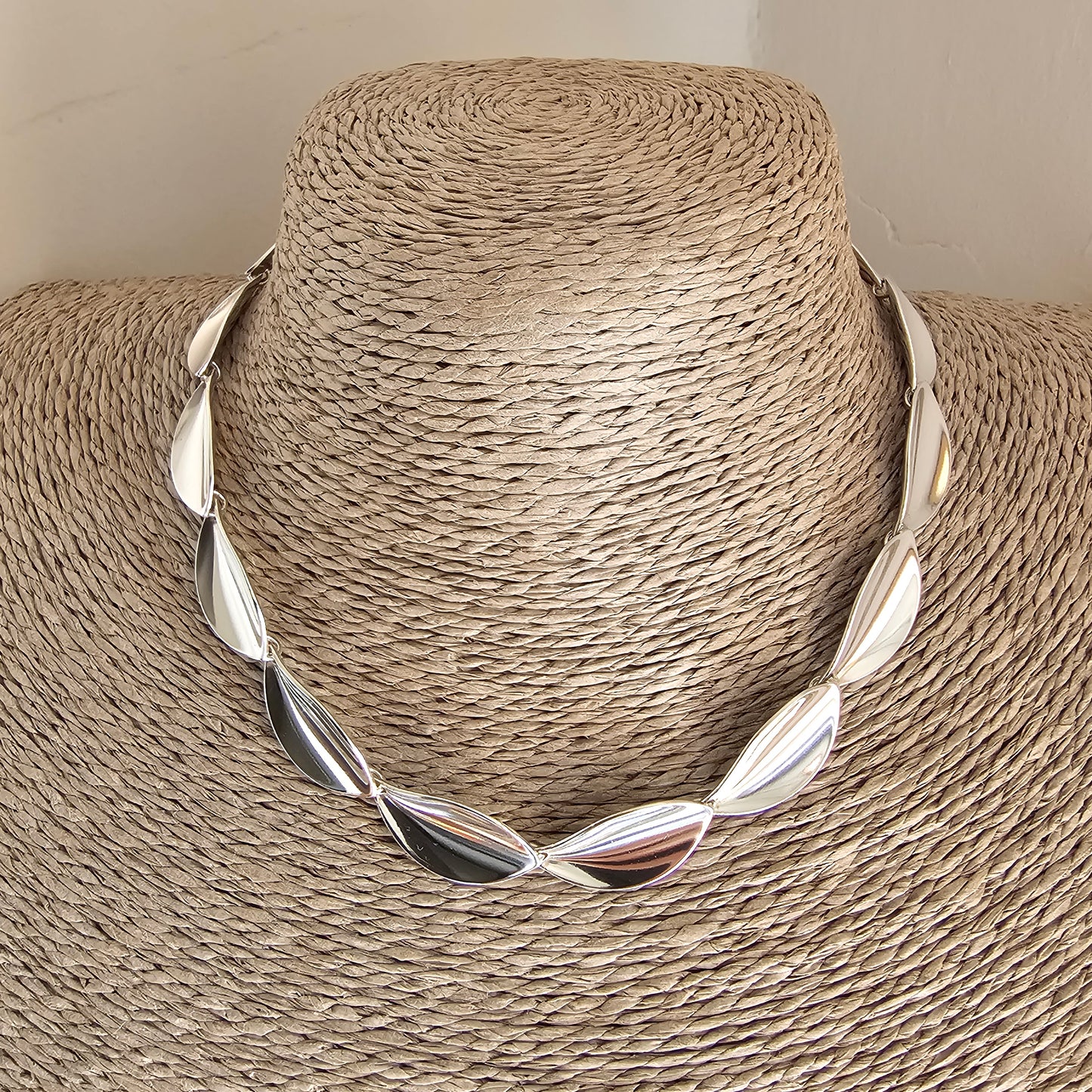 Silver twisted collar necklace with leaf-like segments displayed on a straw hat form.