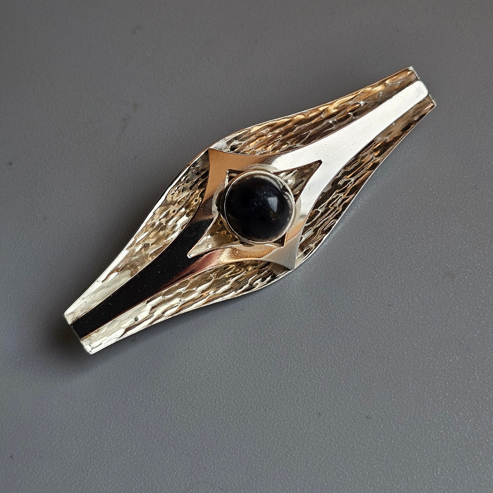 Decorative brooch or pin featuring a central black stone set in a geometric metal design with textured edges.