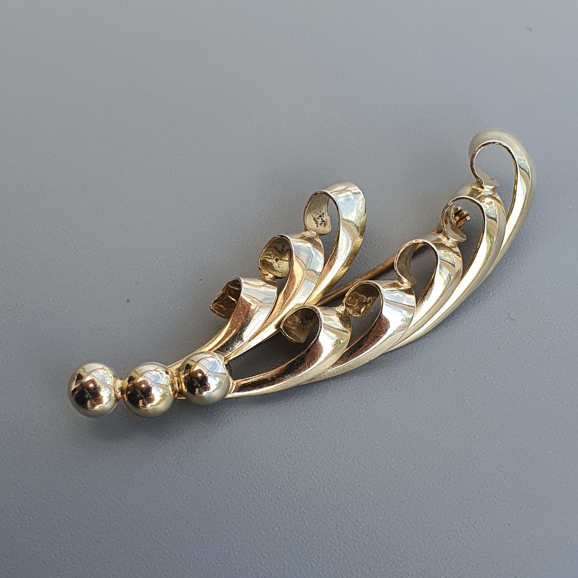 Gold-toned brooch with a curved, leaf-like design featuring small spherical accents.