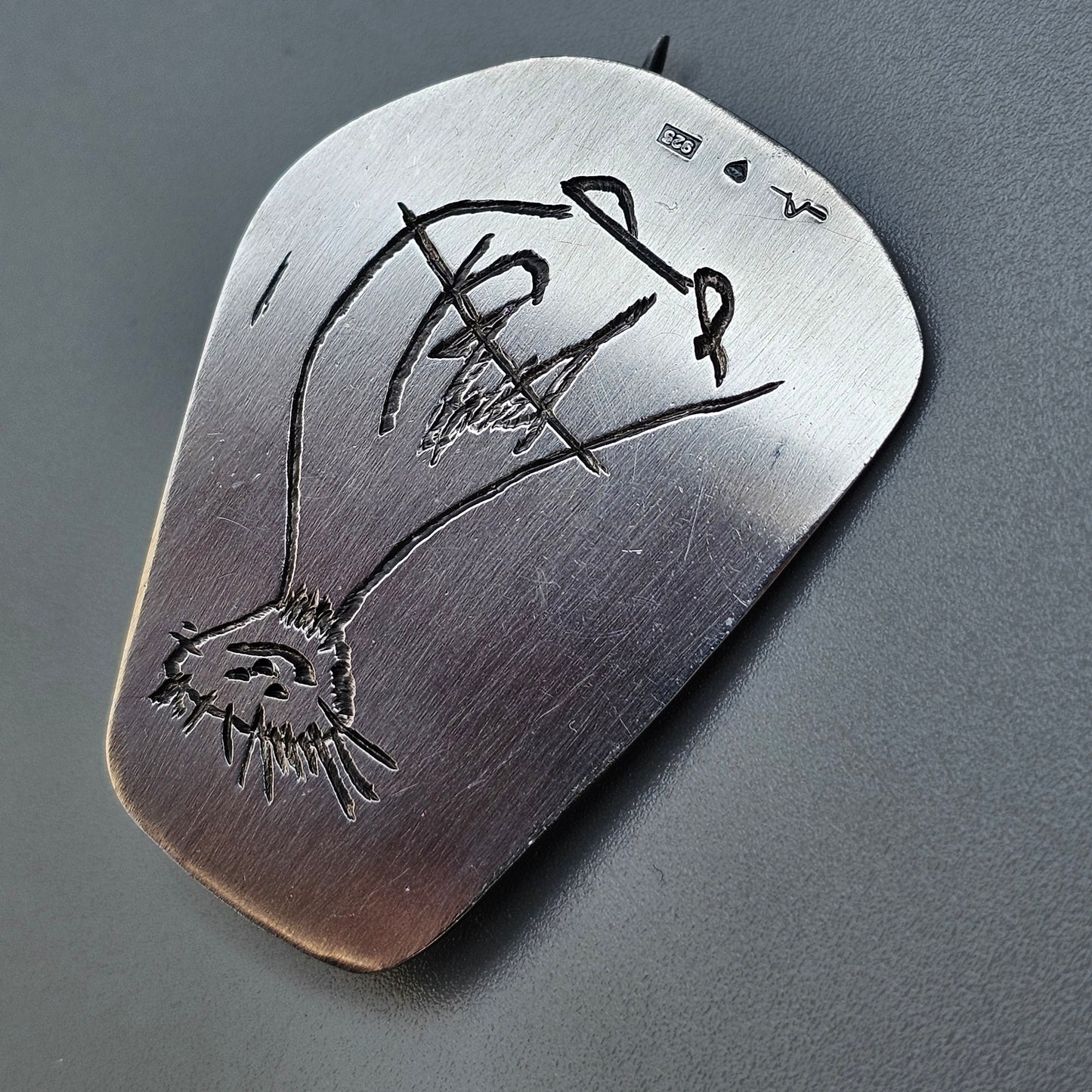 Metal guitar pick with an engraved design resembling a stylized eye and signature.