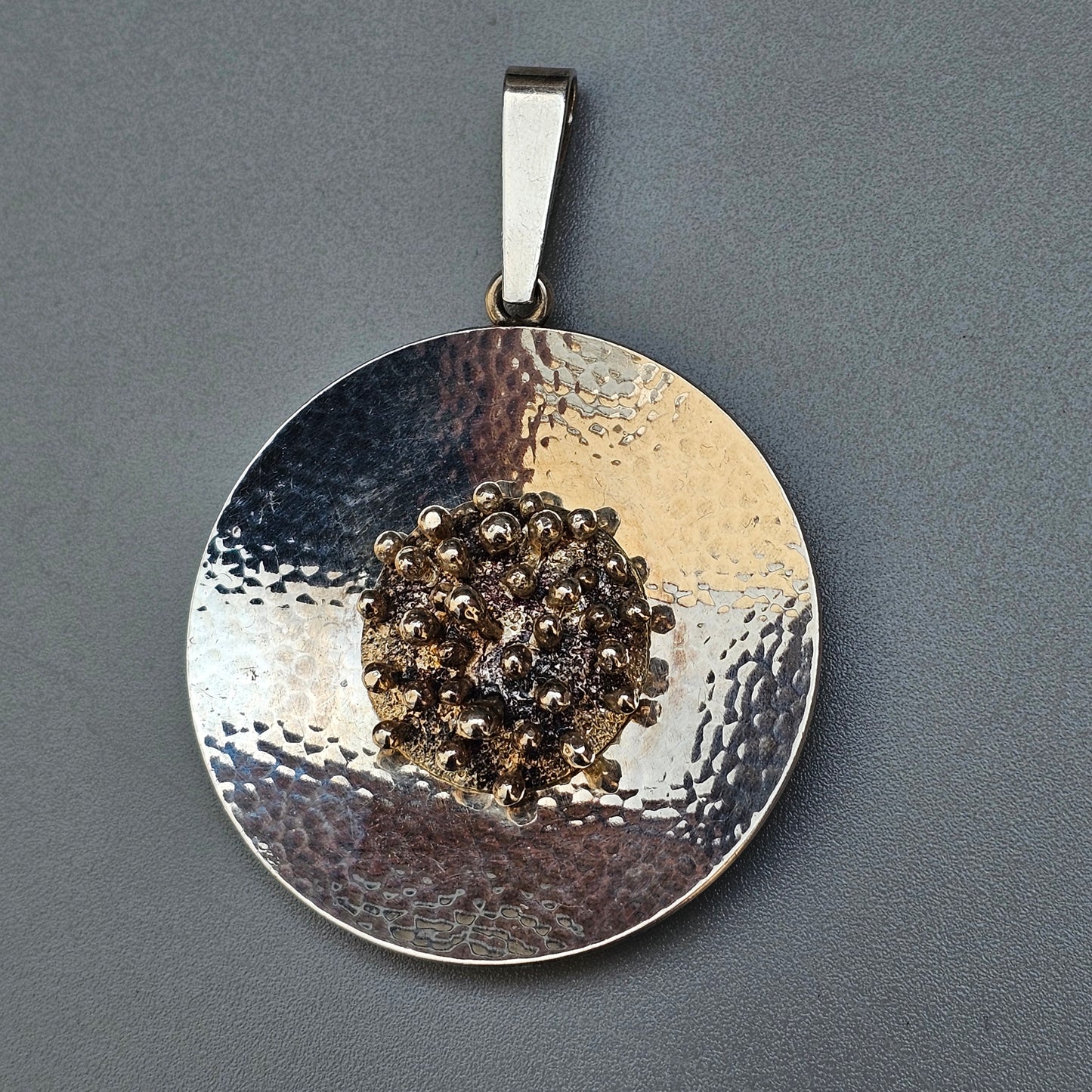 Circular silver pendant with a textured surface and a cluster of small gold-colored spheres in the center.