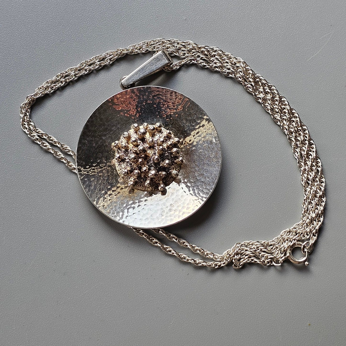 Silver pendant necklace with a textured circular disc and central floral-like design.