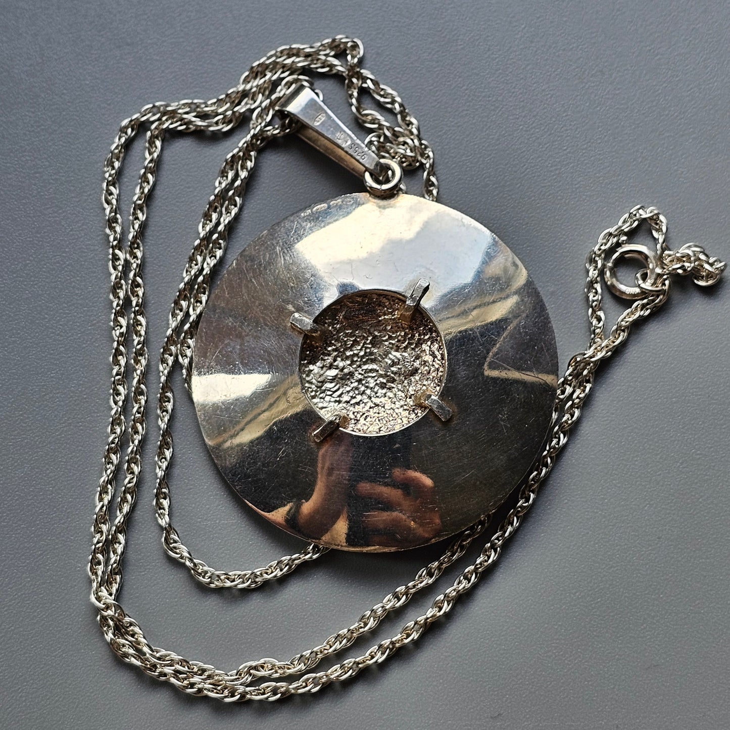 Silver pendant with a circular design and central hole, attached to a chain necklace.