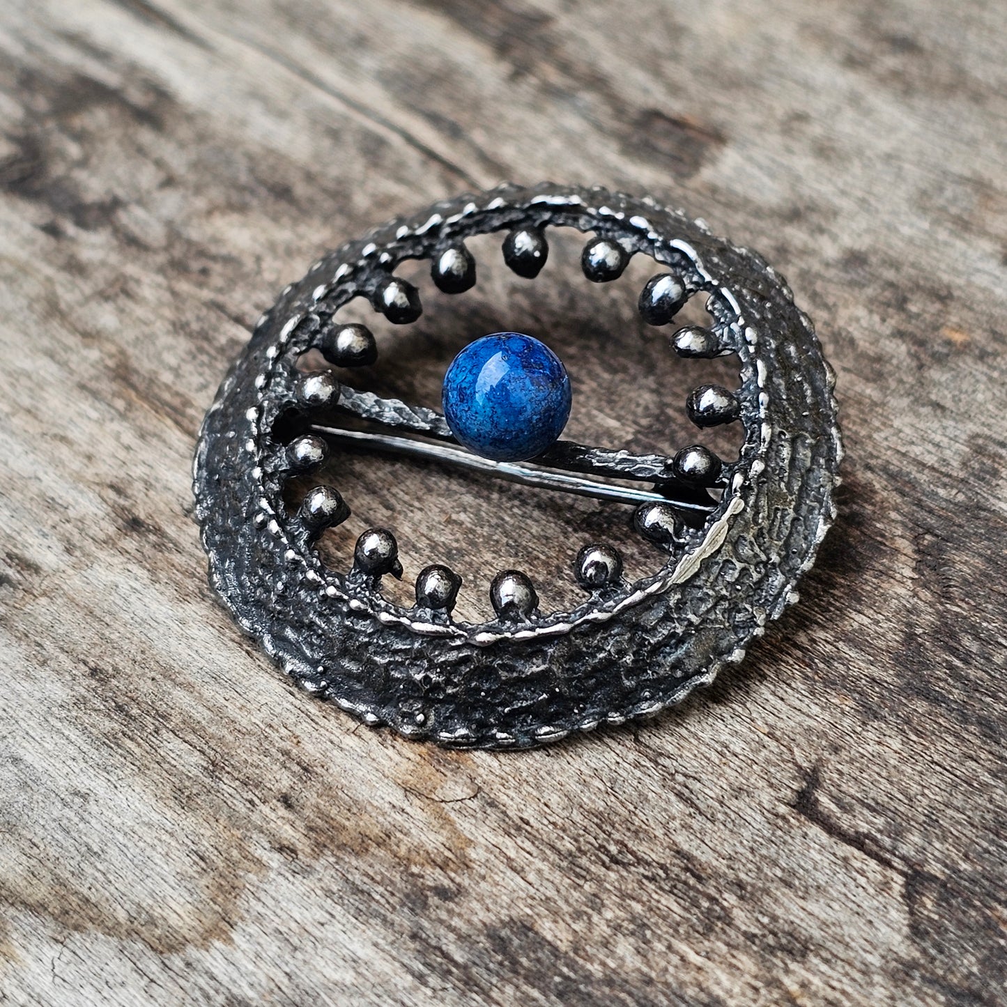 Circular metallic brooch or pendant with a blue bead at its center.