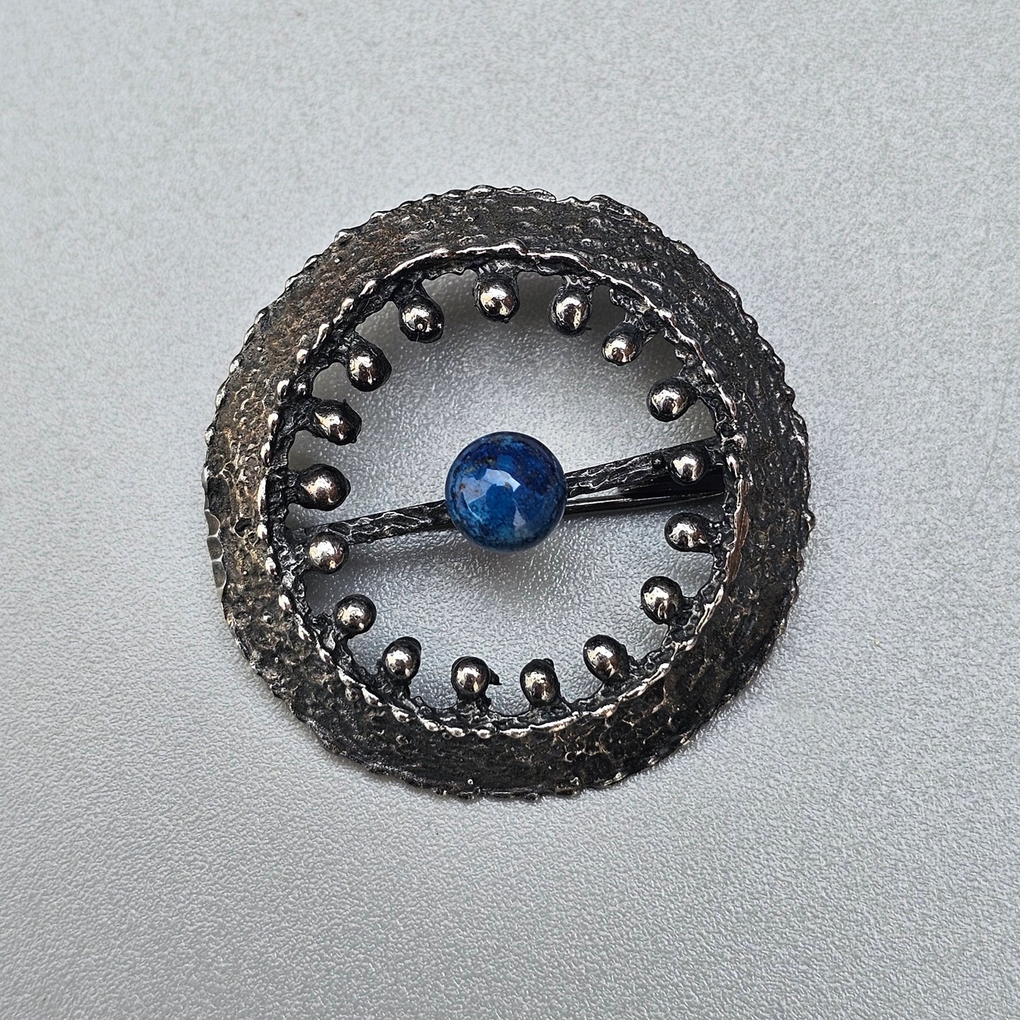 Circular metallic brooch with a blue bead at its center and studded edges.