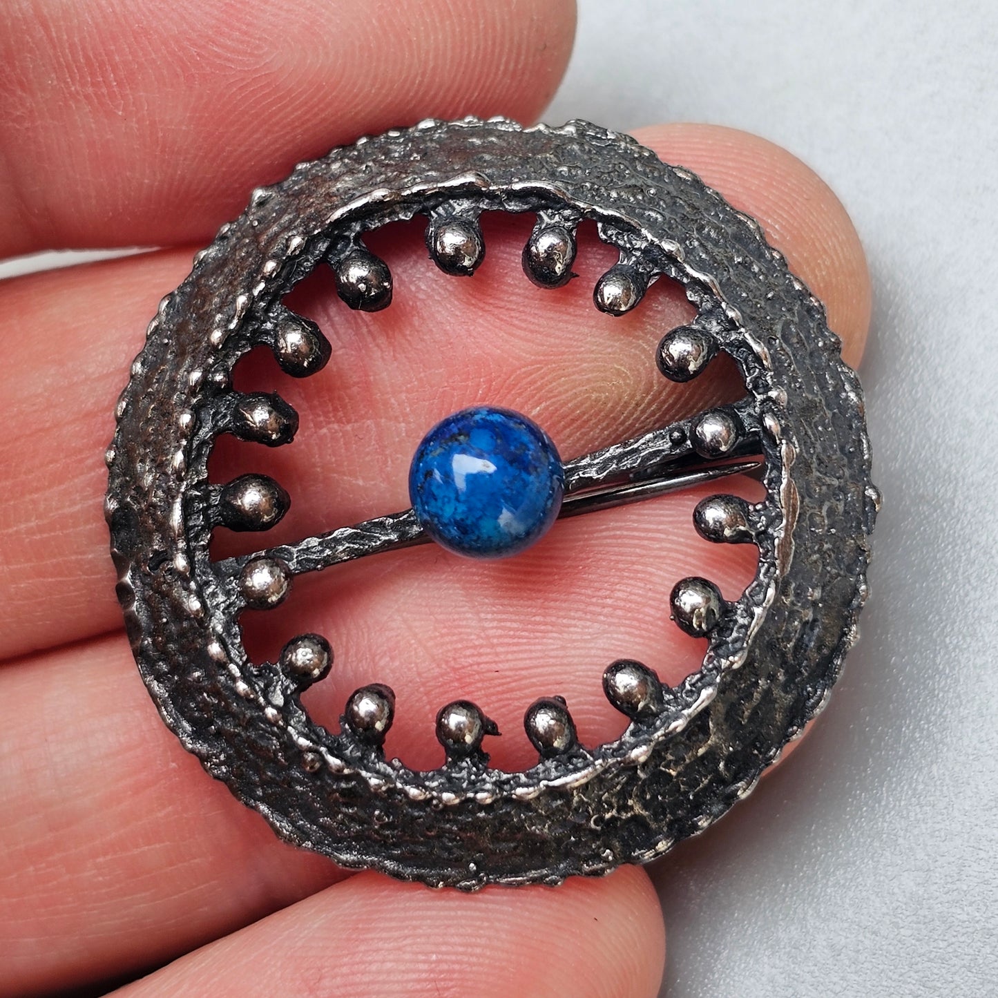 Circular metallic brooch or pendant with a blue gemstone center and beaded border.