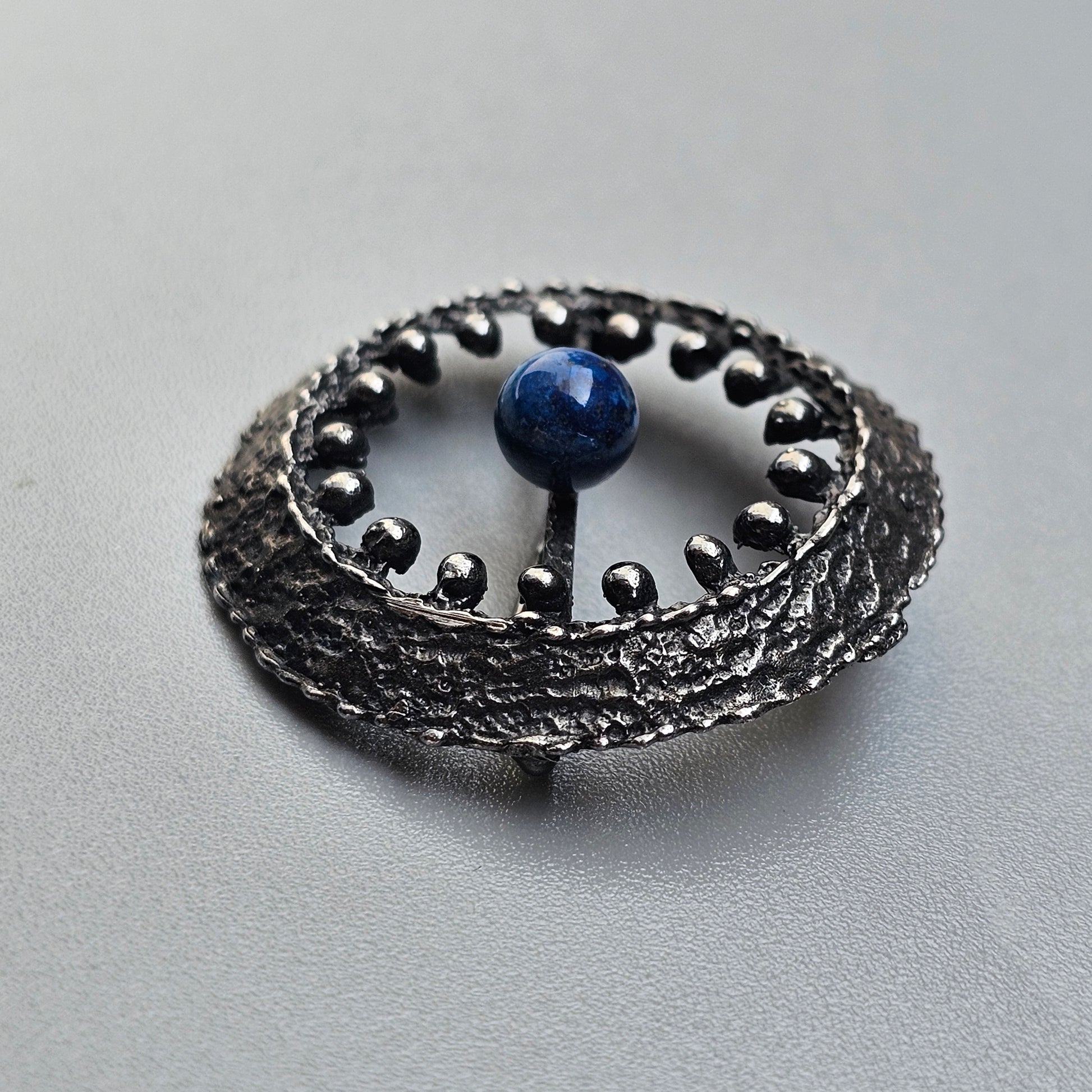 Circular metallic brooch or pendant with a blue stone at its center, surrounded by decorative silver beads.