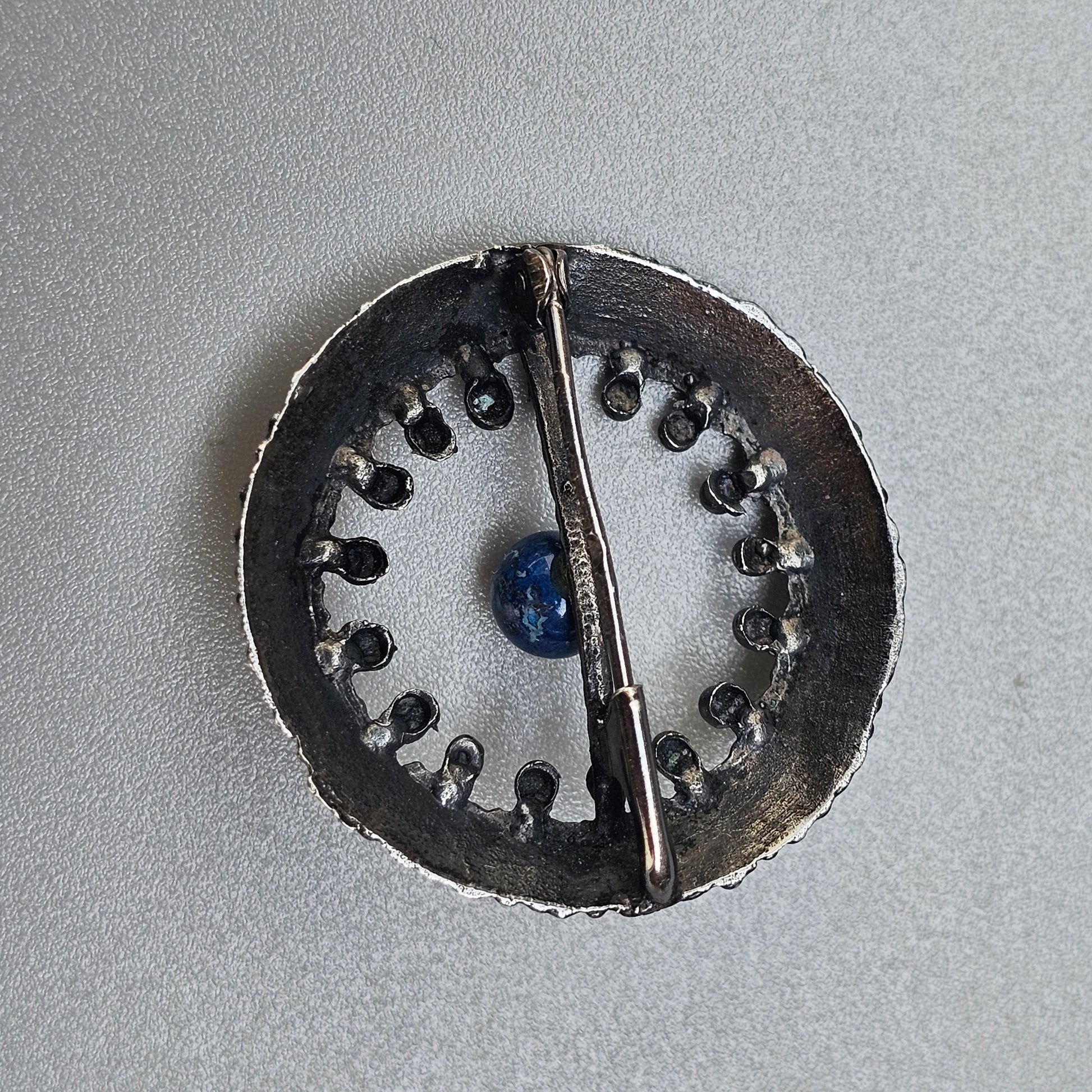 Circular metal brooch or pendant with a central blue stone and decorative perforations around the edge.