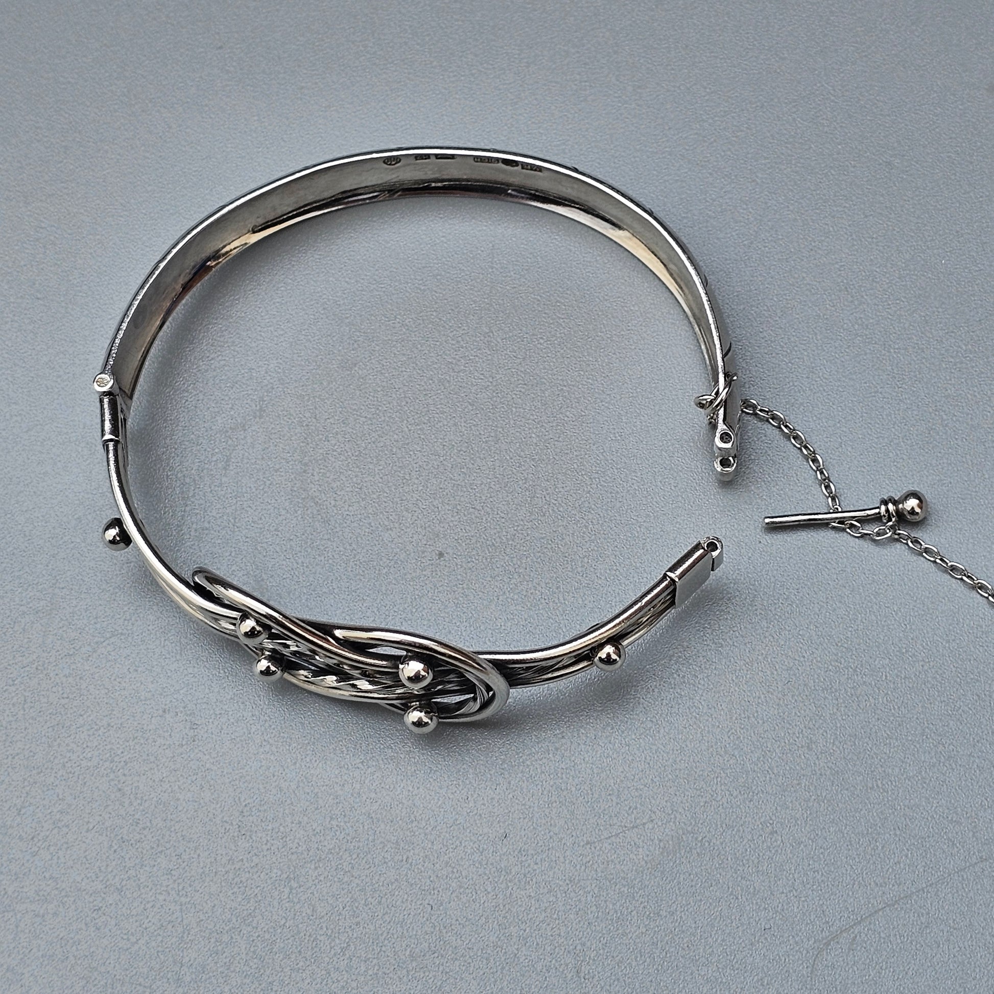 Silver bangle bracelet with an intricate floral or leaf design and a safety chain.