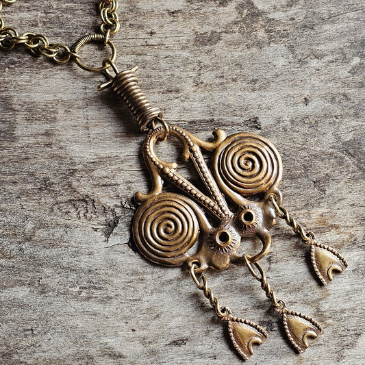 Ornate brass pendant with spiral designs and dangling elements on a chain.