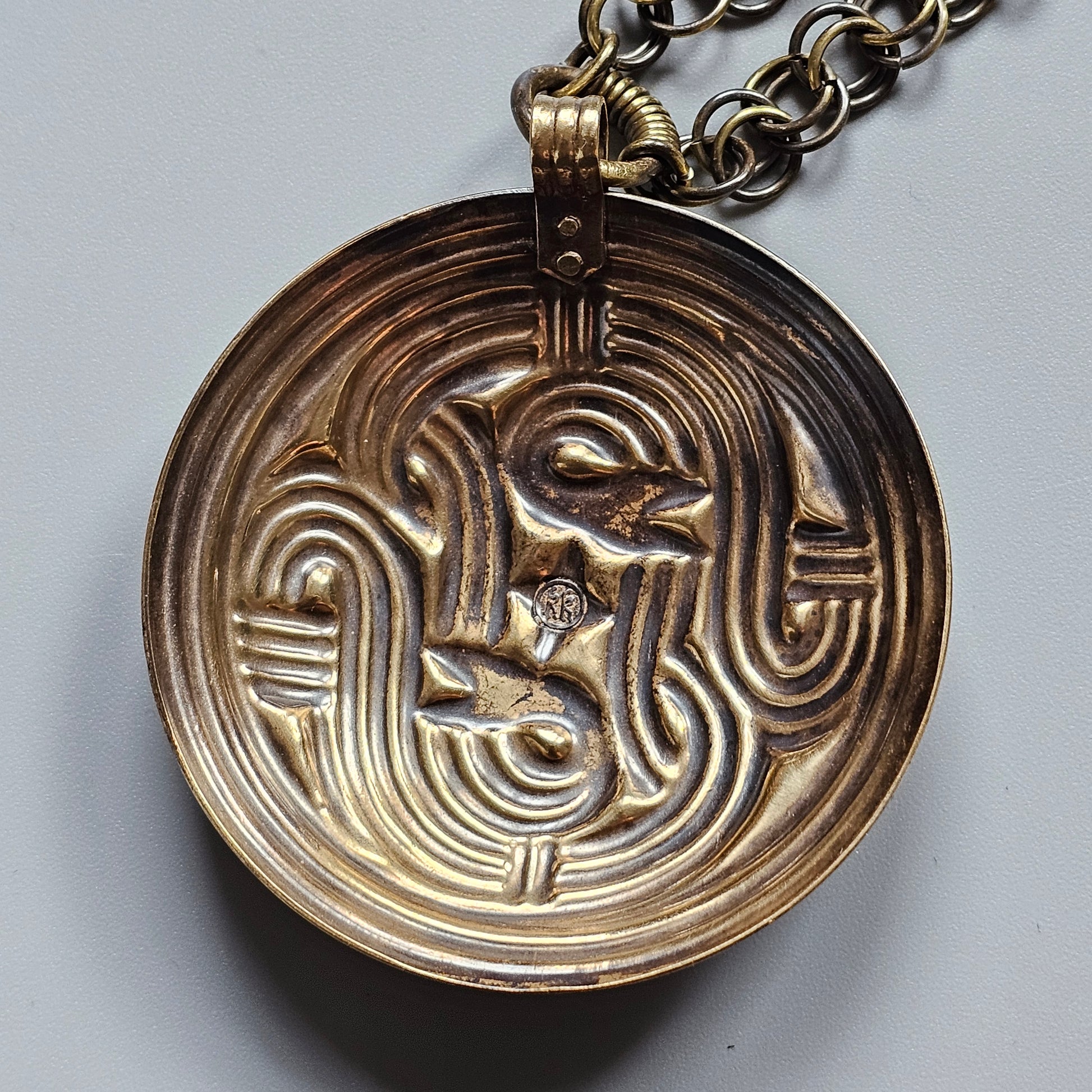 Circular bronze pendant with an intricate abstract face design on a chain.