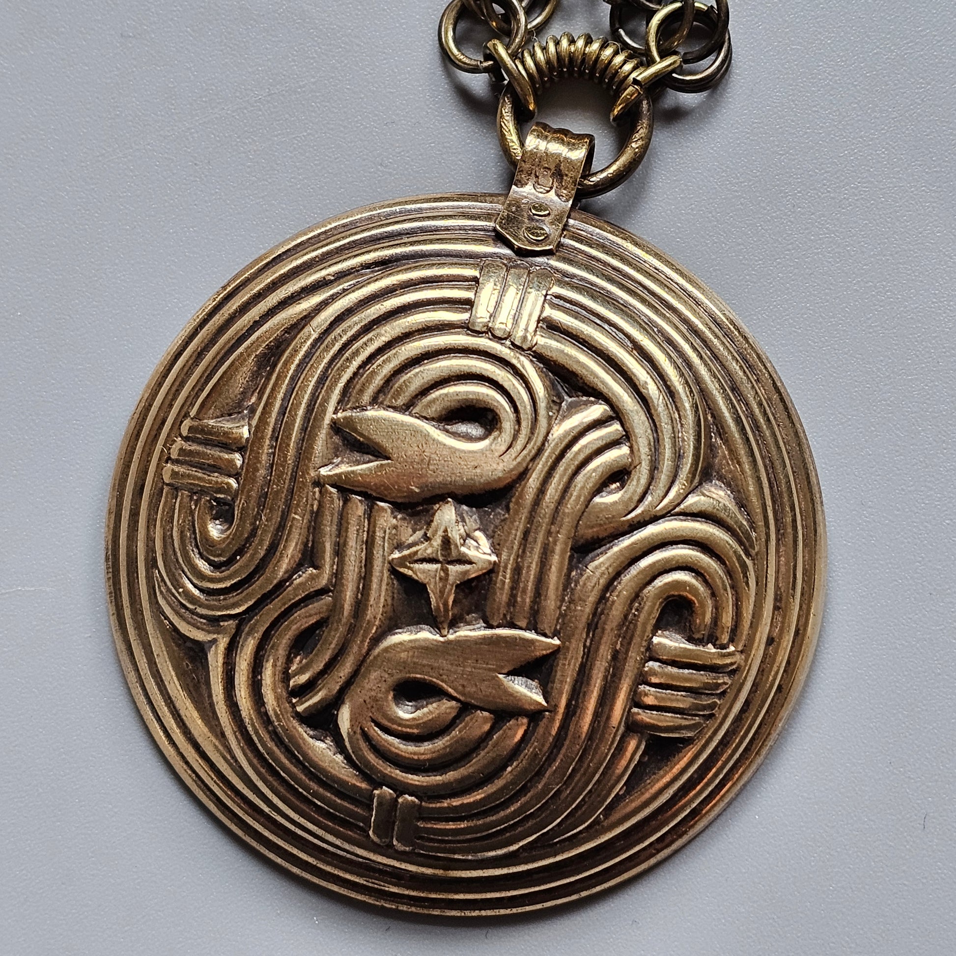 Circular gold-toned pendant with an intricate abstract design featuring swirling patterns and stylized animal shapes.