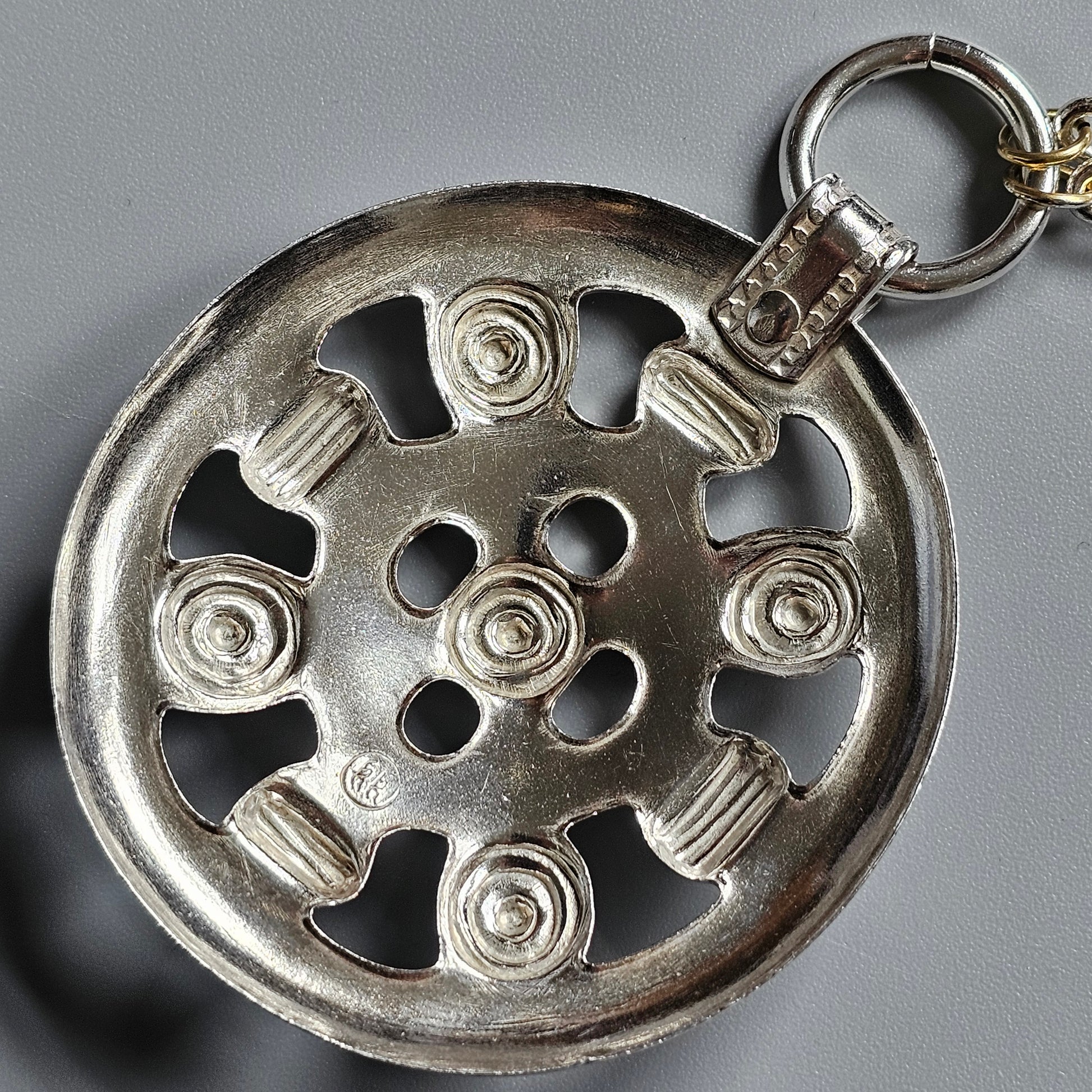 Circular metal keychain or pendant with an ornate wheel-like design featuring spirals and columns.