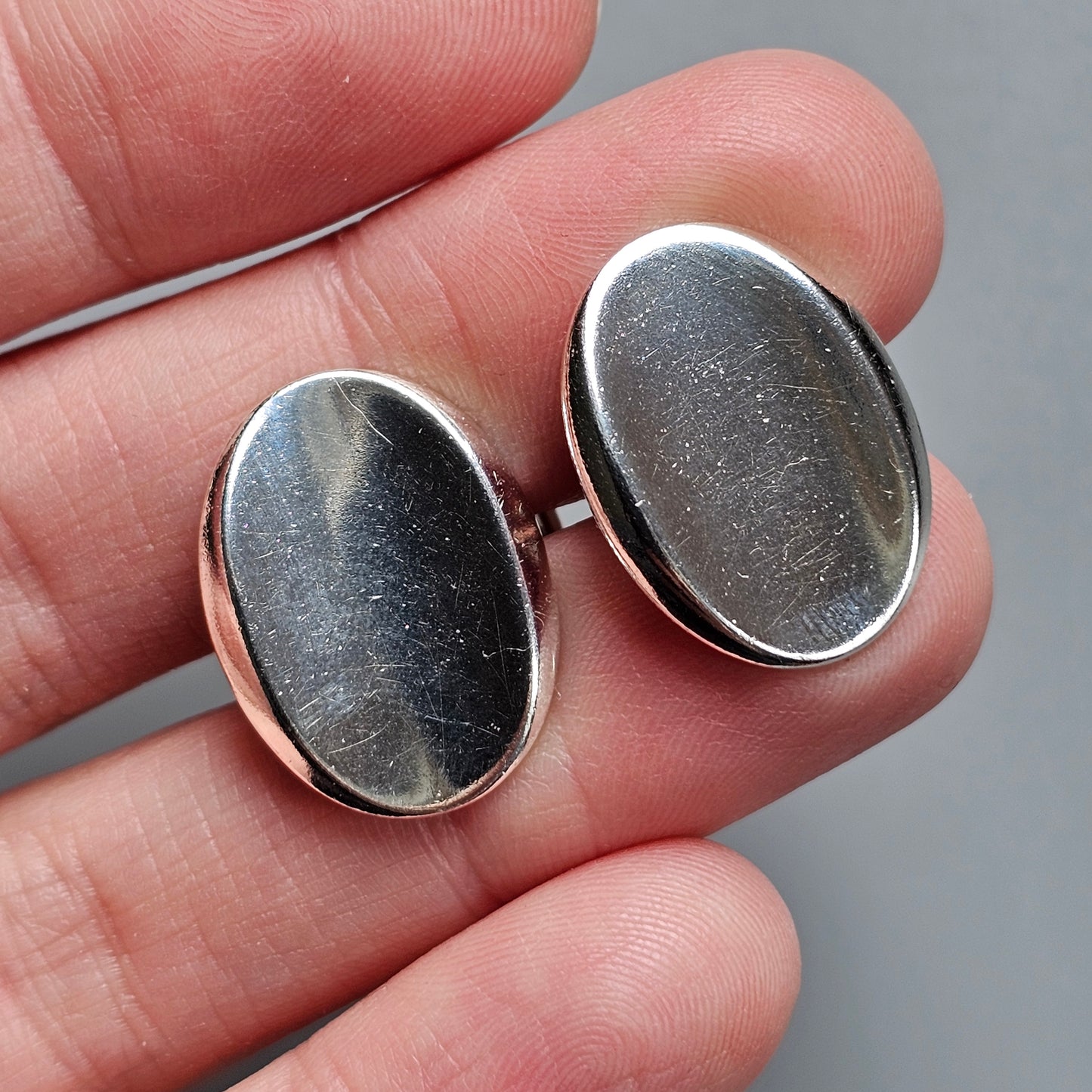 Pair of oval silver cufflinks held between fingers.