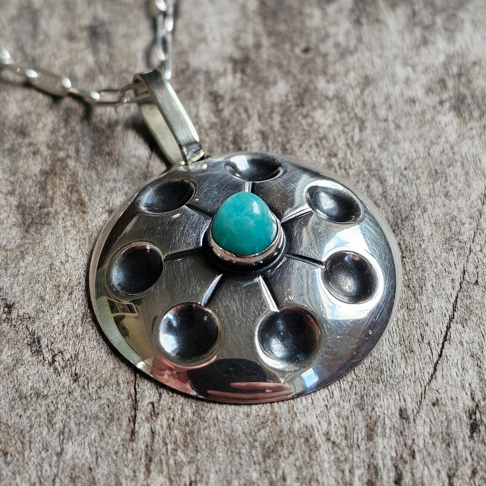 Silver pendant necklace with a circular design featuring a central turquoise stone surrounded by small indentations.