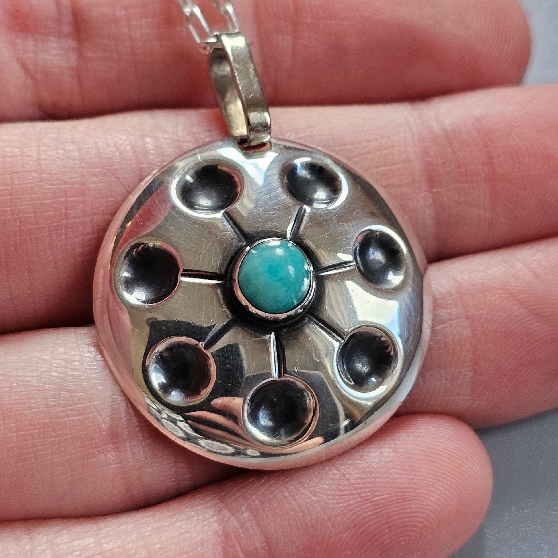 Silver pendant with a circular design featuring a central turquoise stone surrounded by smaller dark indentations.