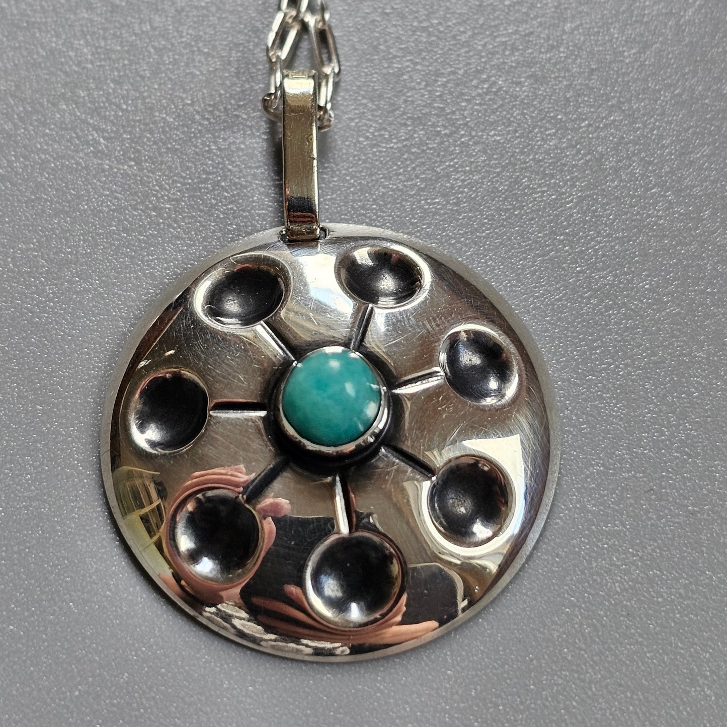 Silver pendant with a circular design featuring a central turquoise stone surrounded by smaller circular indentations.