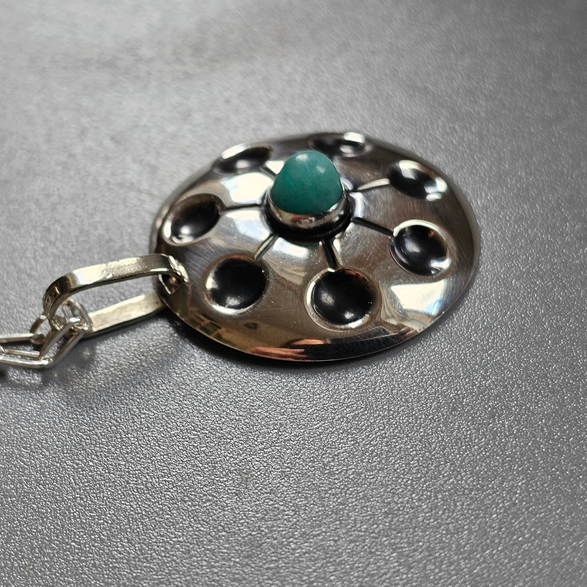 Silver pendant with a circular design featuring a central turquoise stone and surrounding holes.