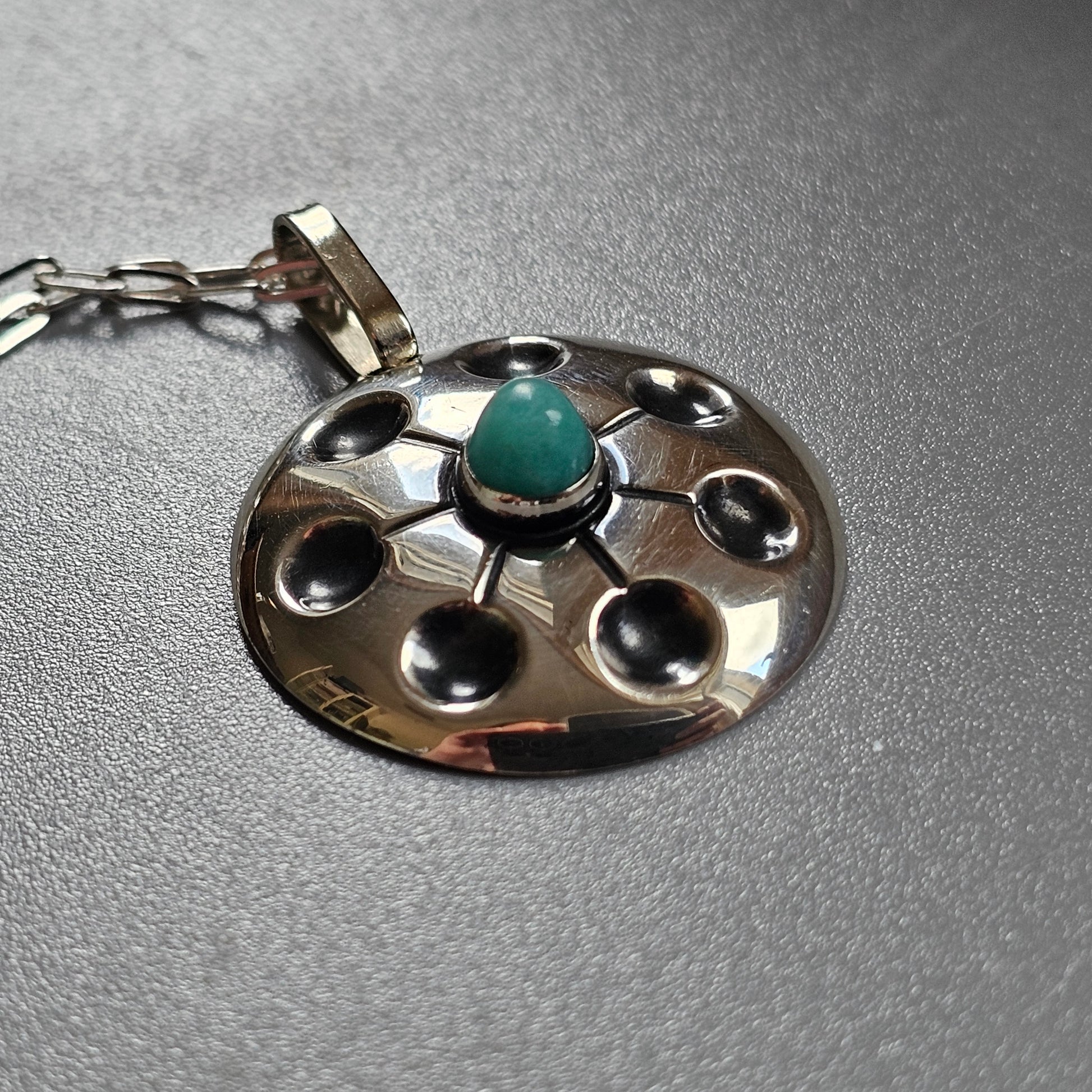 Silver pendant with a circular design featuring a central turquoise stone surrounded by indentations.