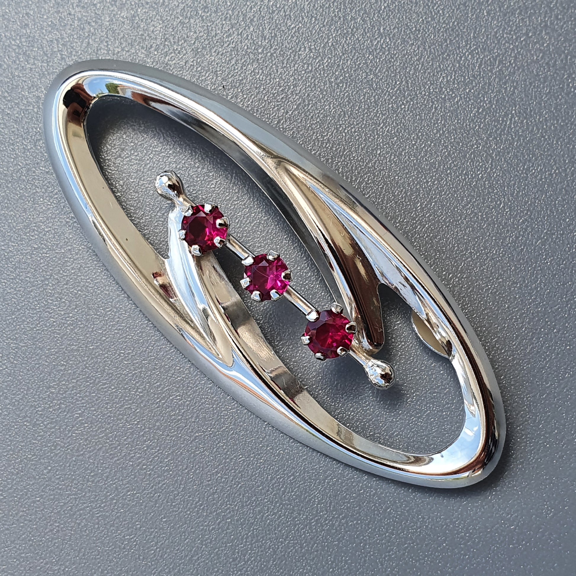 Oval-shaped silver brooch with three red gemstones set in a curved design.