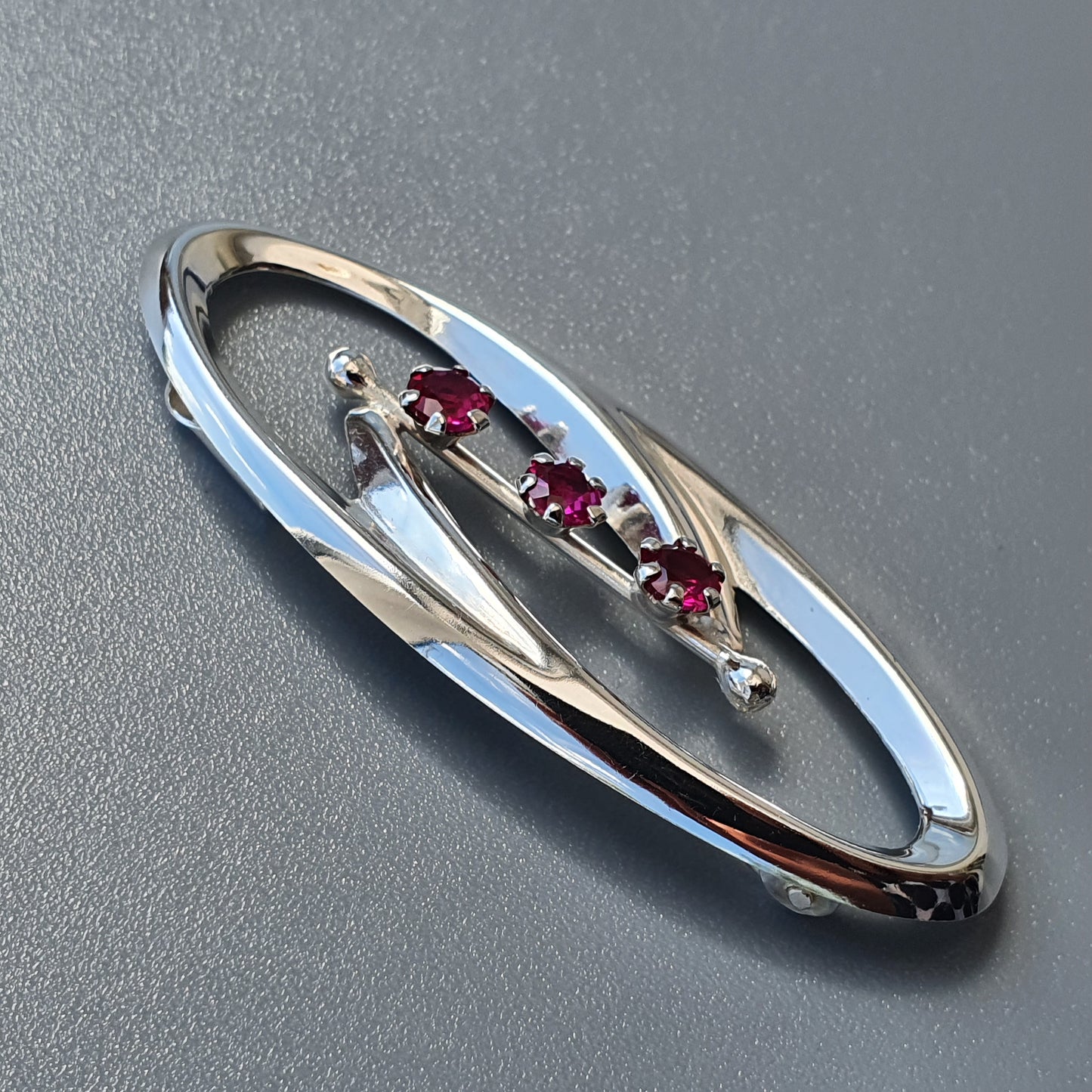 Oval-shaped silver brooch with three small red gemstones set in the center.