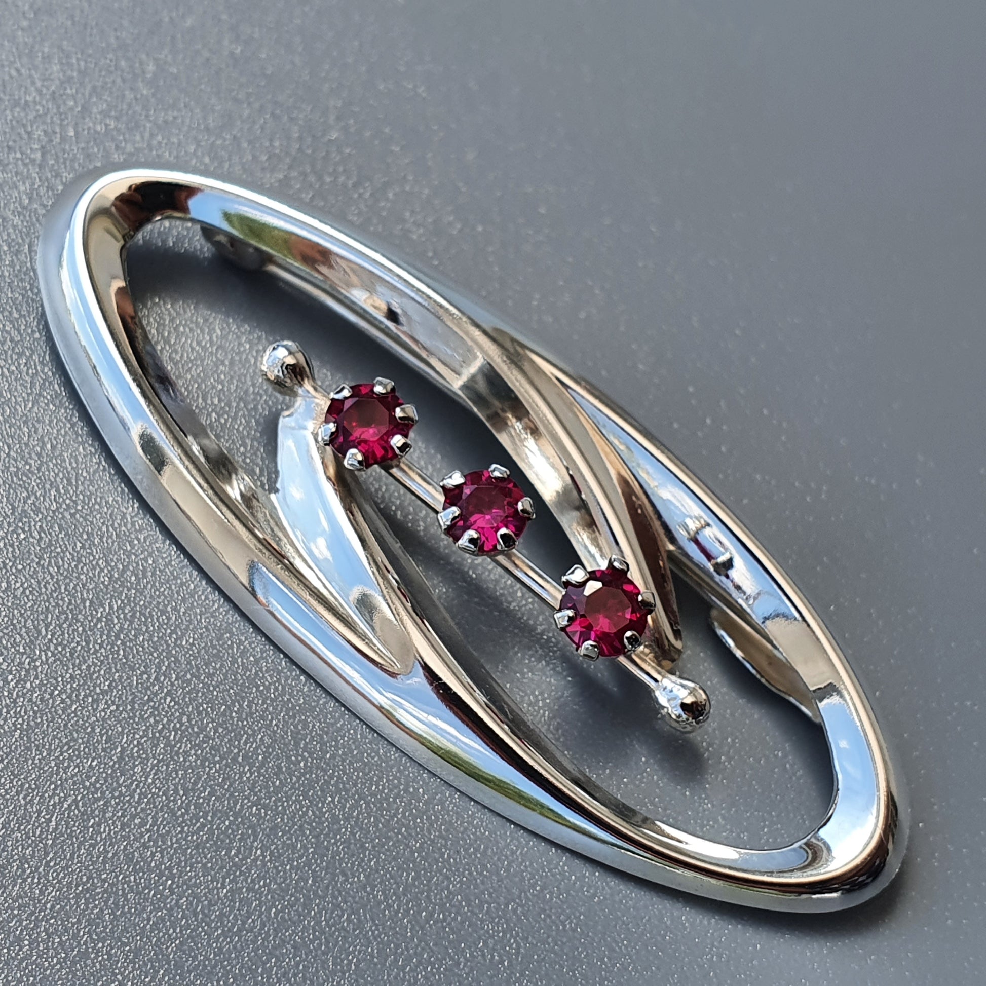 Oval-shaped silver brooch with three red gemstones set diagonally across its center.