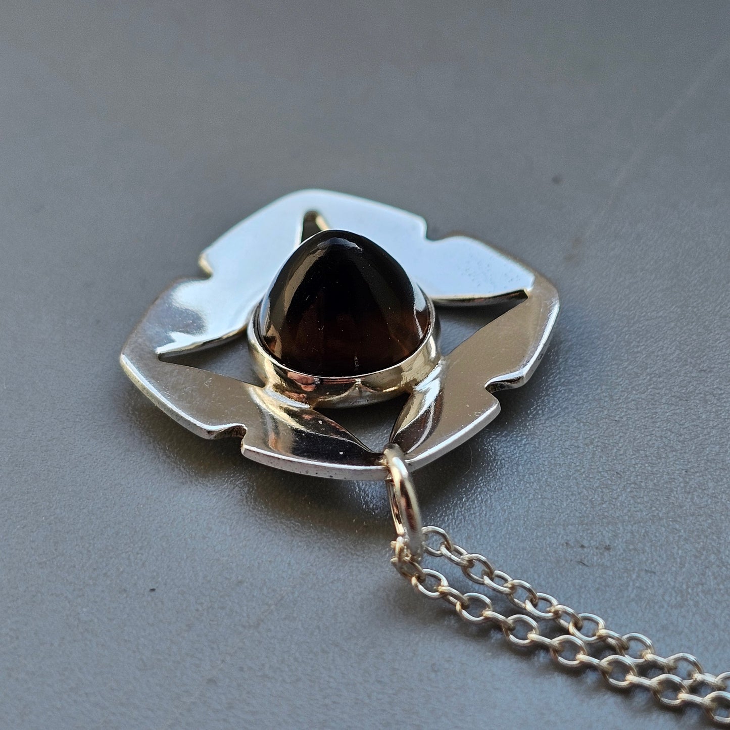 Silver pendant with a dark gemstone set in a stylized flower or cross-shaped design, attached to a chain.