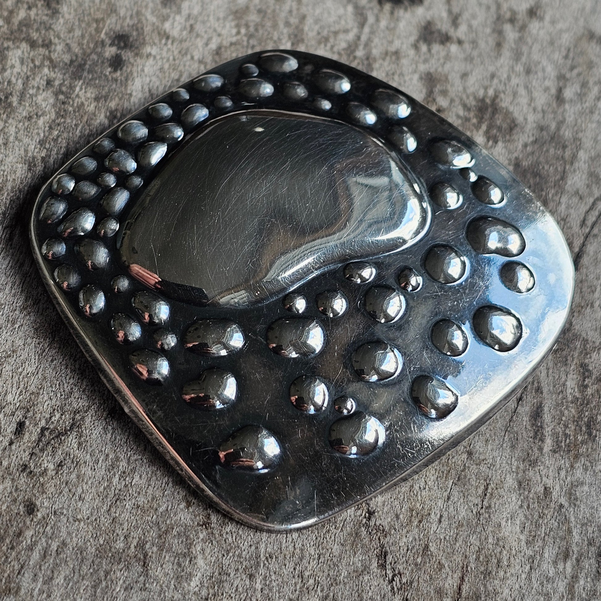 Metallic pendant or brooch with a smooth center surrounded by raised bubble-like dots.