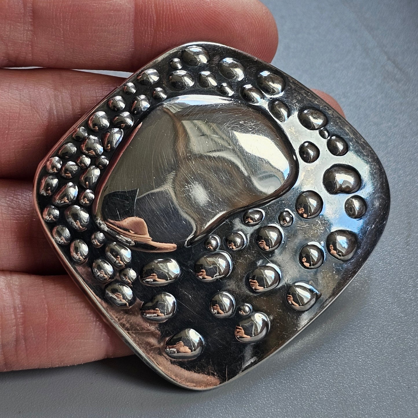 Metallic pendant or brooch with a smooth central area surrounded by a textured border of raised dots.