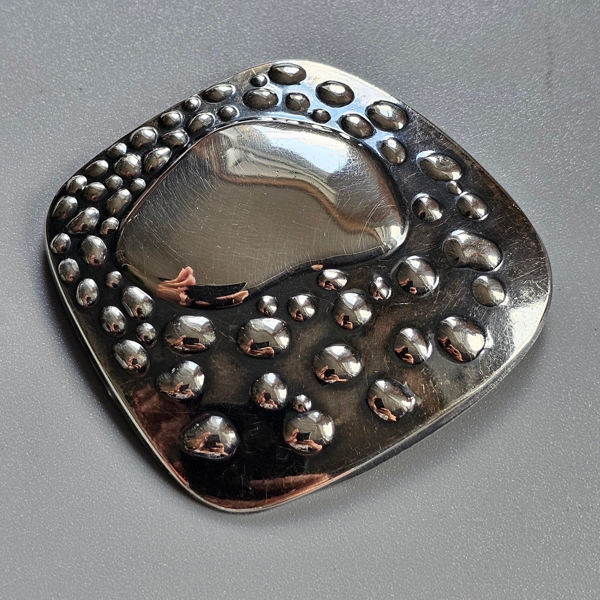 Metallic brooch or pendant with a rounded square shape and raised bubble-like decorations.