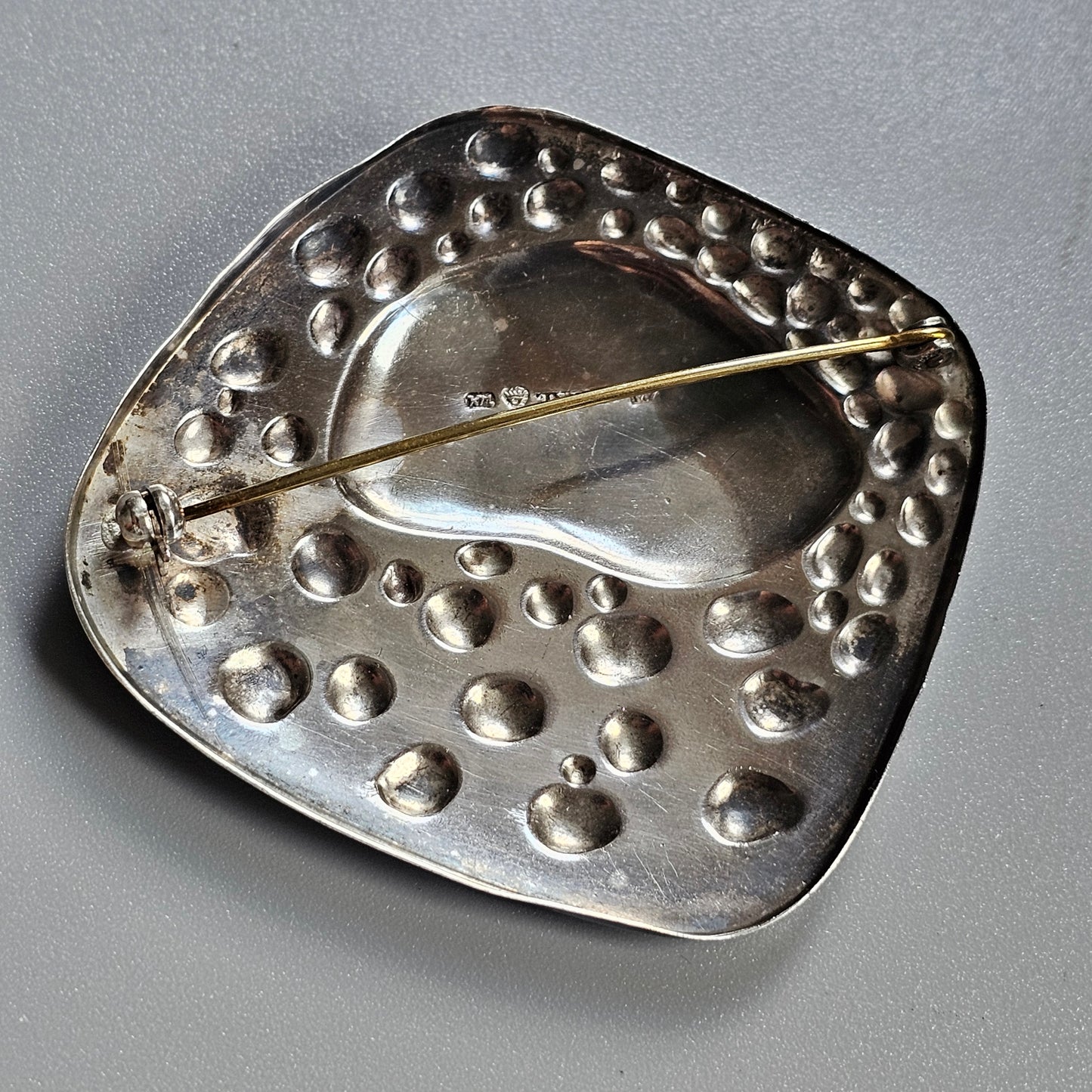 Metallic brooch or pin with a dimpled surface and a long needle-like fastener.