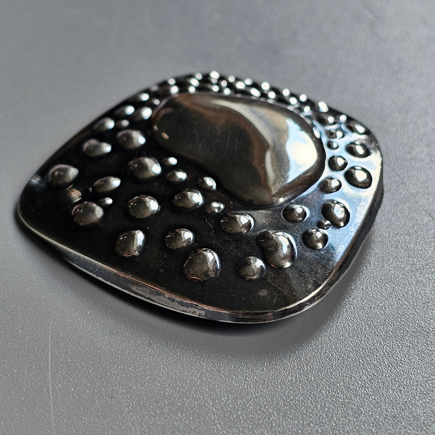 Metallic brooch or pendant with a rounded square shape and dotted pattern.