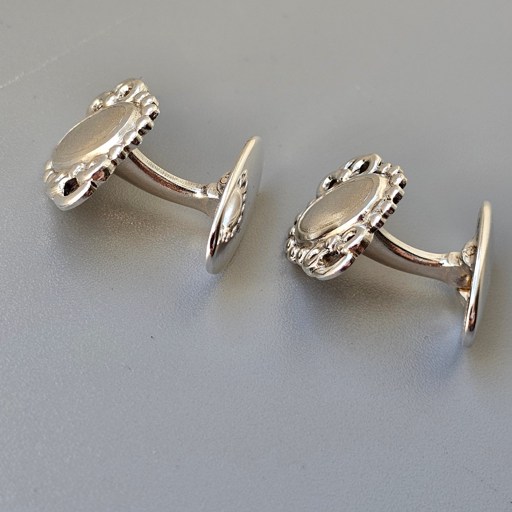 Pair of silver oval-shaped cufflinks with decorative edges.