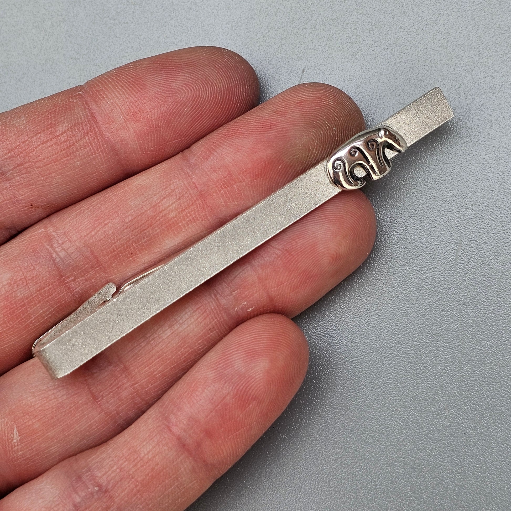 Silver tie clip with decorative scroll design at one end.