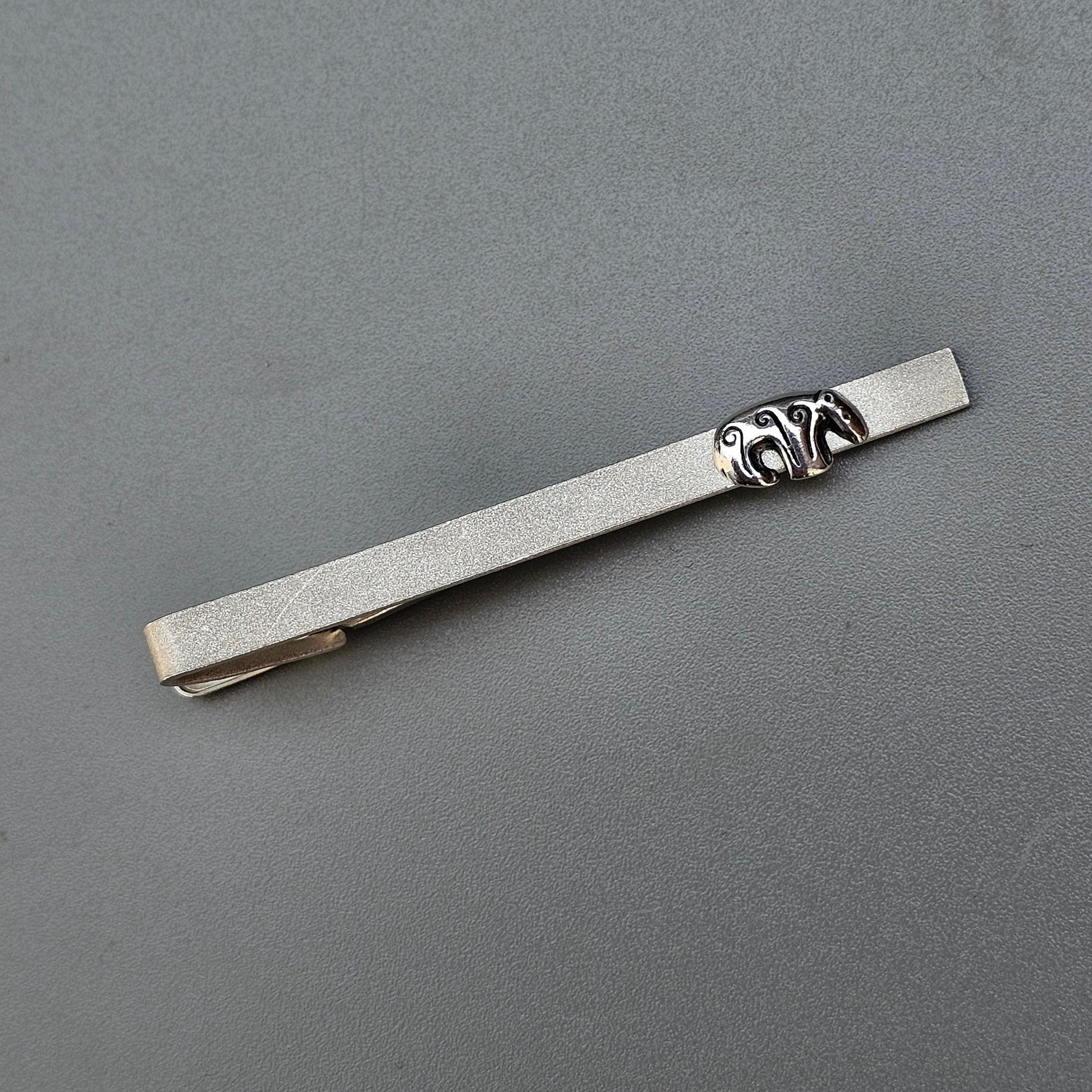 Silver tie clip with an ornate decorative element near one end.