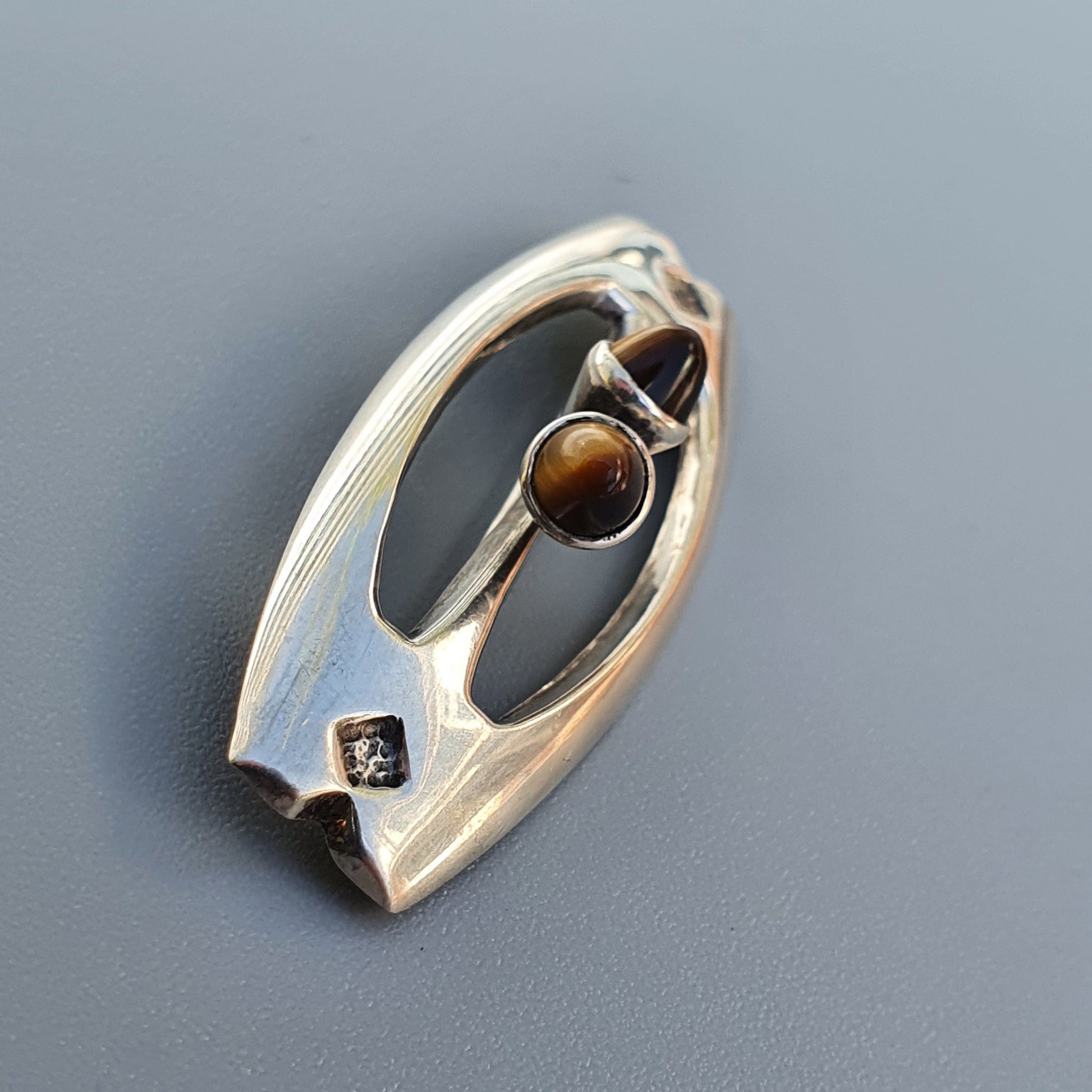 Oval-shaped silver brooch with a tiger’s eye gemstone set in an abstract design.