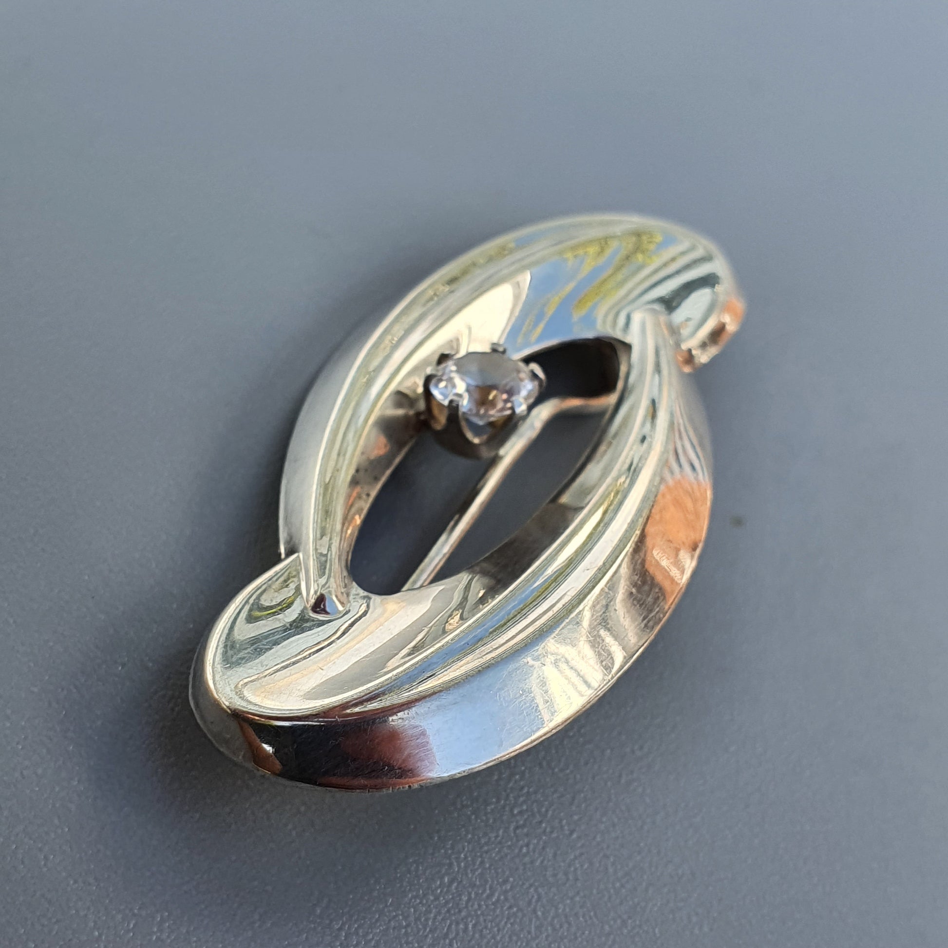 Oval-shaped silver brooch or pin with an Art Nouveau-inspired flowing design and a small gemstone.